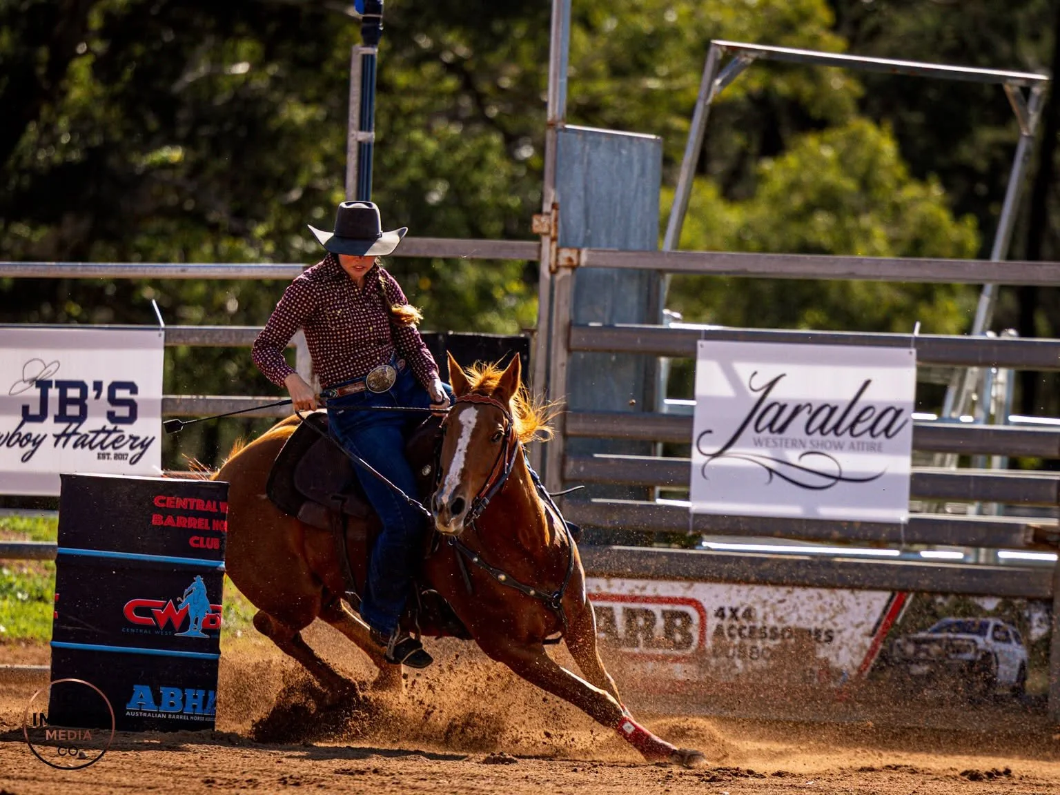 Barrel racer rides a horse around a barrel in a dirt arena.
