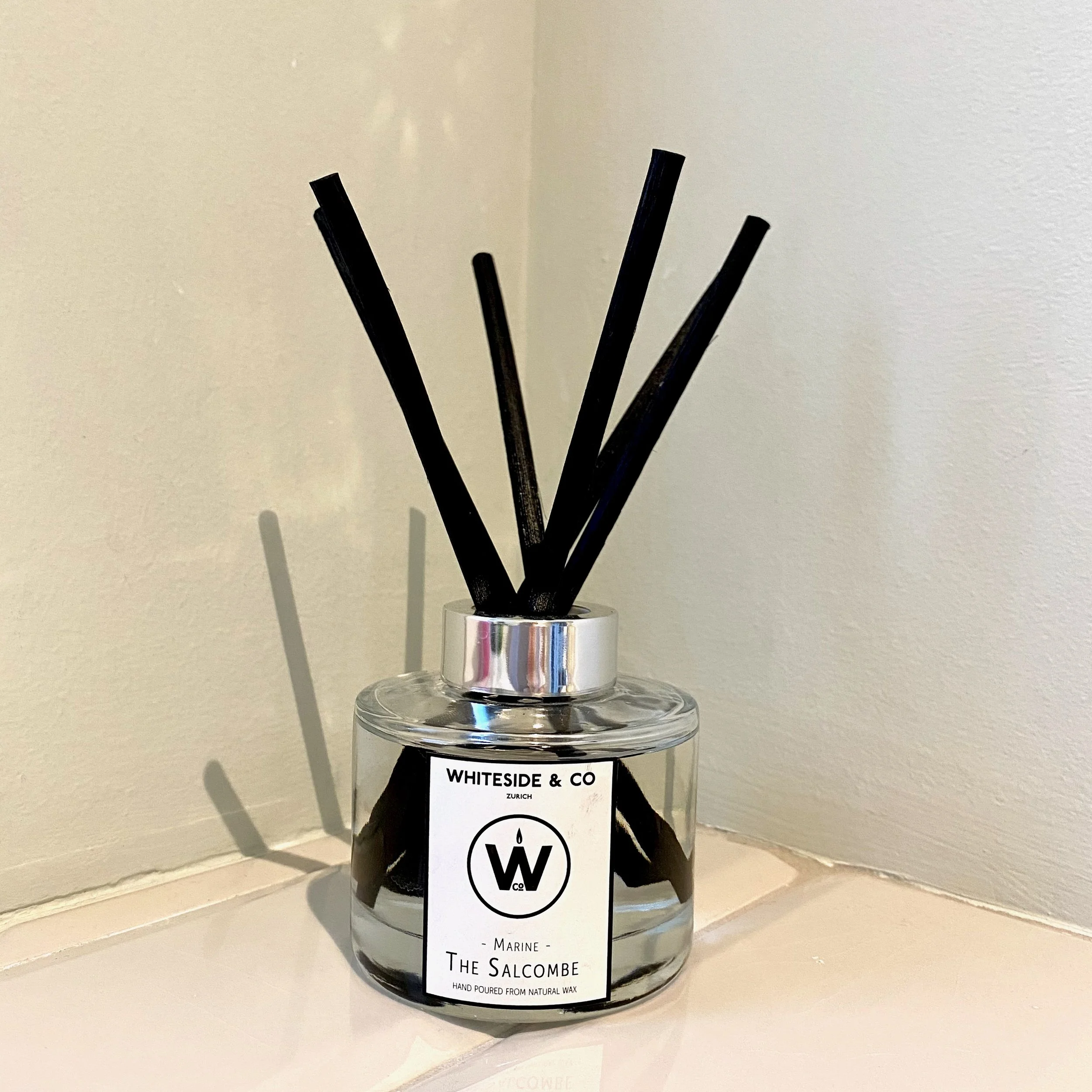A reed diffuser in a clear round bottle with a chrome lid and thick black bamboo reeds