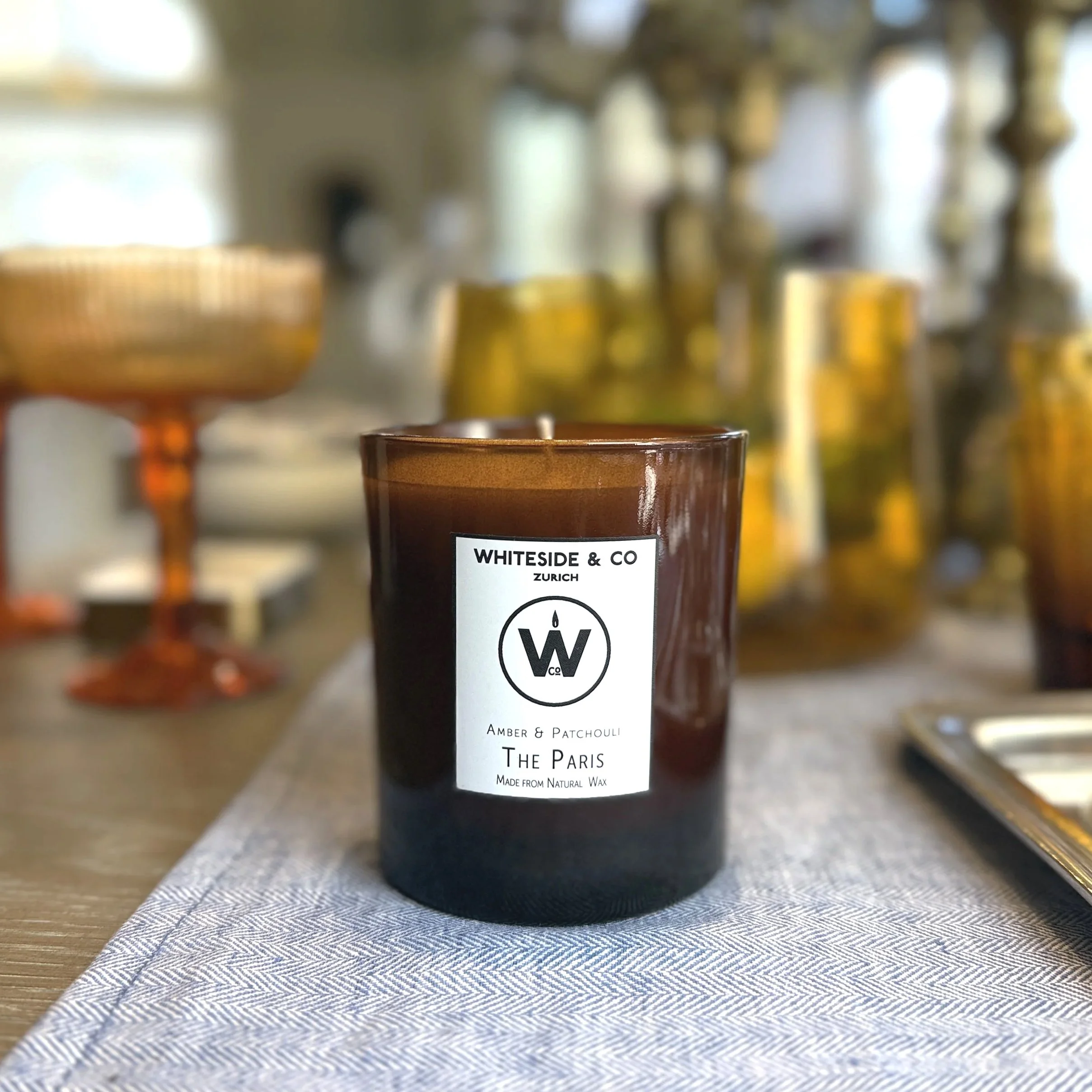 A brown candle with a white label reading 'WHITESIDE & CO ZURICH Amber & Patchouli THE PARIS Made from natural wax' on a table with a white and blue striped tablecloth. In the background are blurred glassware and decorative items.