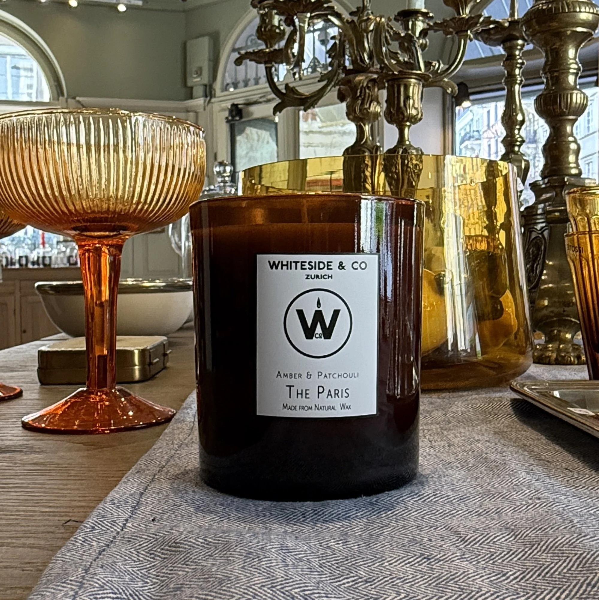 A candle in an amber glass on a decadent table with a champagne glass and candelabra behind it