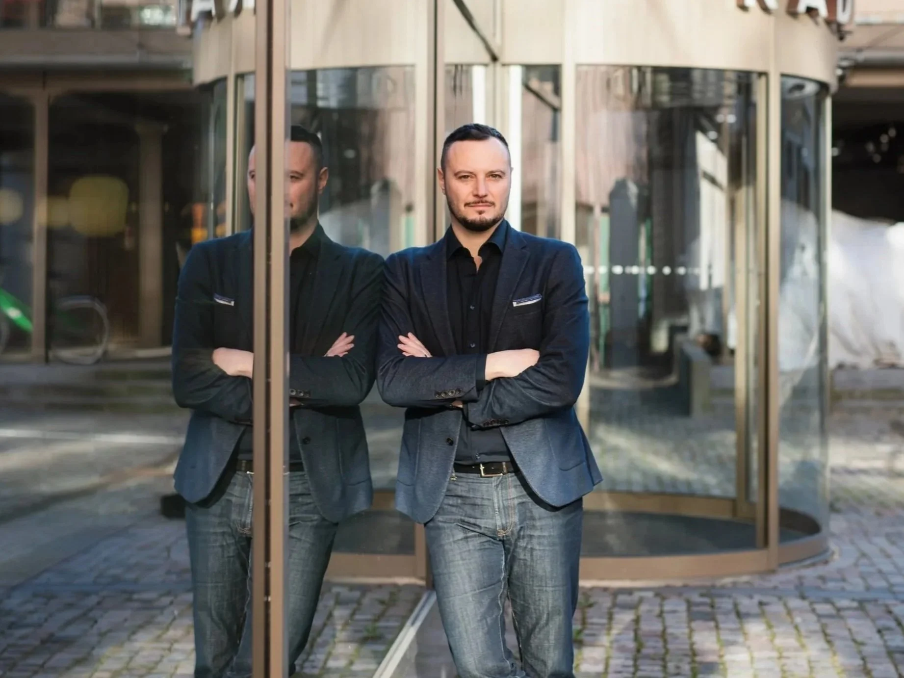A man in a blue blazer and jeans stands with arms crossed, reflected in glass doors with a round building behind him.