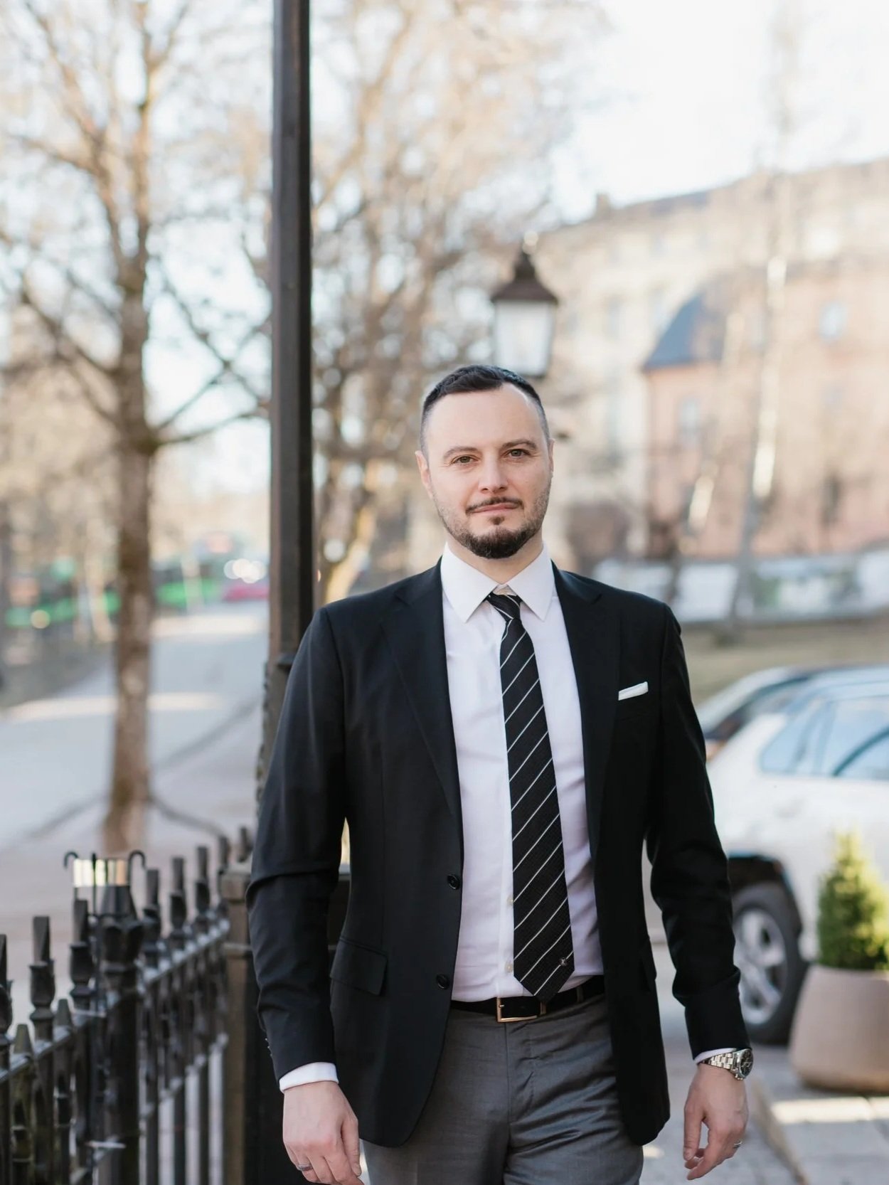 A man in a suit standing outdoors on a city street with trees and parked cars in the background.