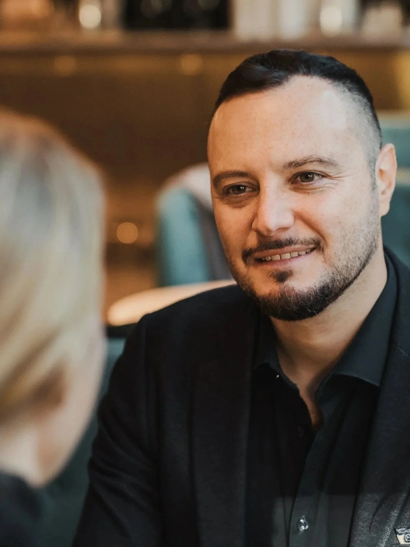 A man with short dark hair and a beard smiling, wearing a black shirt, in a warmly lit indoor setting, possibly a restaurant or cafe.