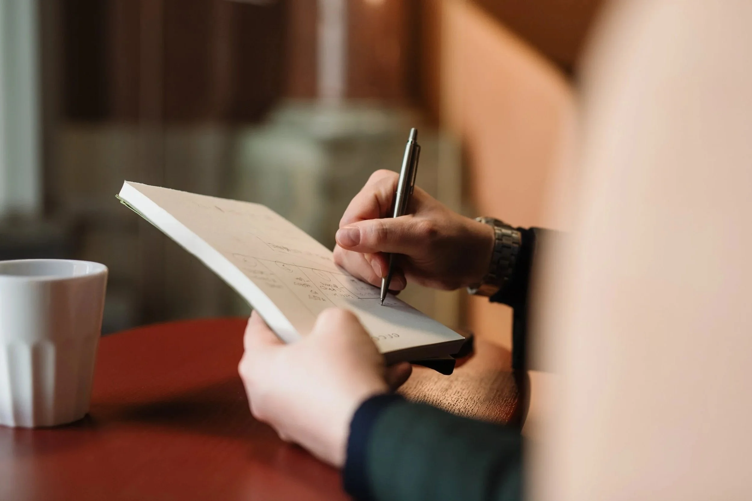 Close-up of a person writing in a notebook with a black pen, sitting at a wooden table in a cozy indoor setting. Two white coffee cups are on the table.