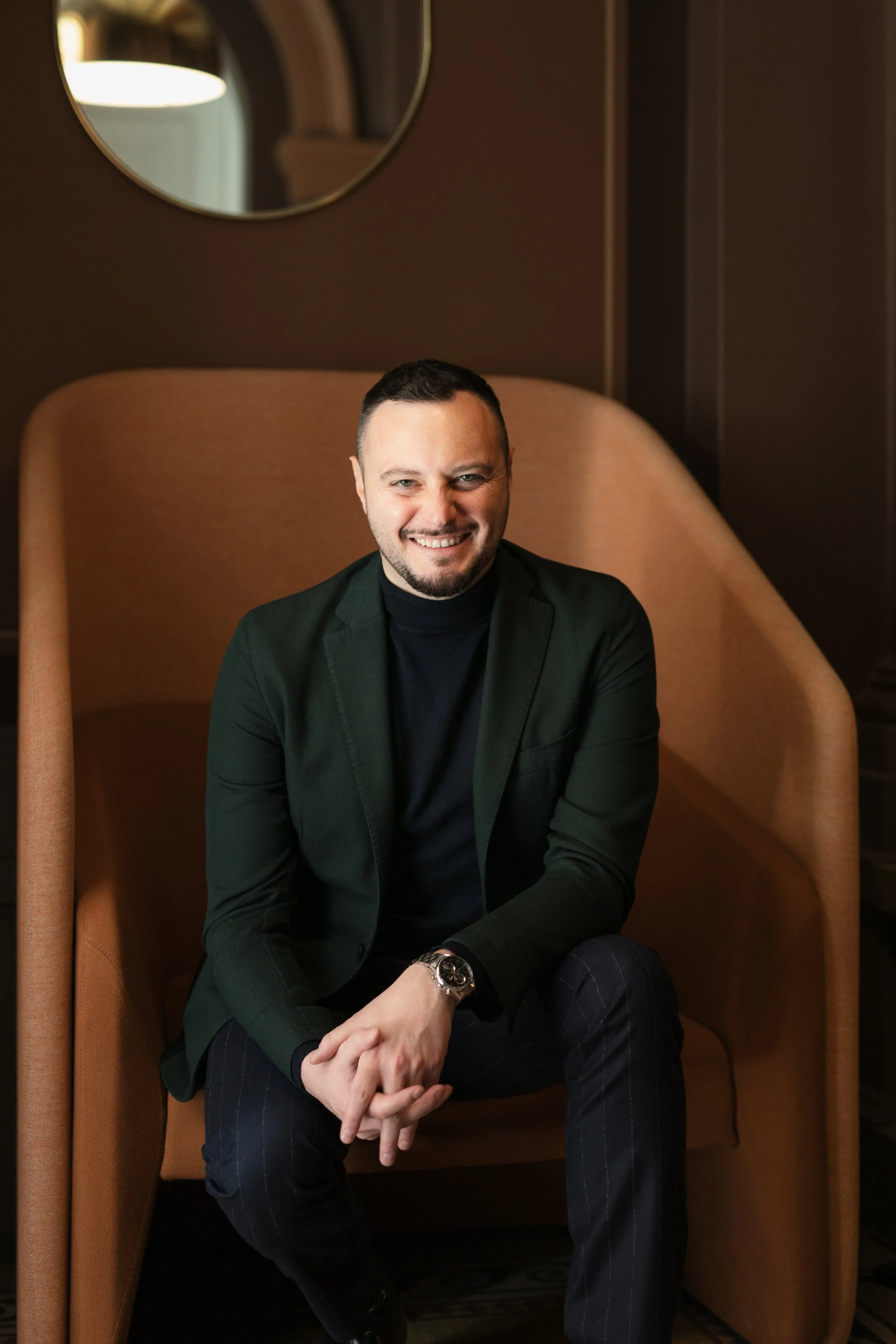 A smiling man wearing a black blazer and black turtleneck sits in a modern, high-backed beige chair in a warmly lit room.