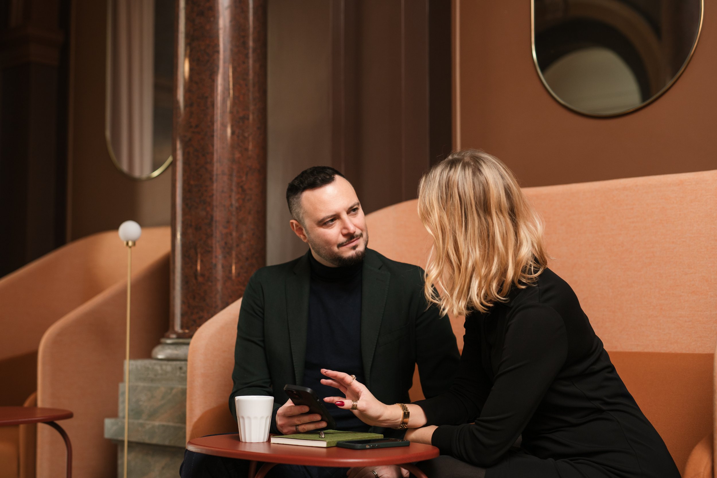 A man and a woman sitting at a small table in a restaurant, looking at a smartphone together, with cups and a notebook on the table, in a warmly lit interior with round mirrors on the wall.