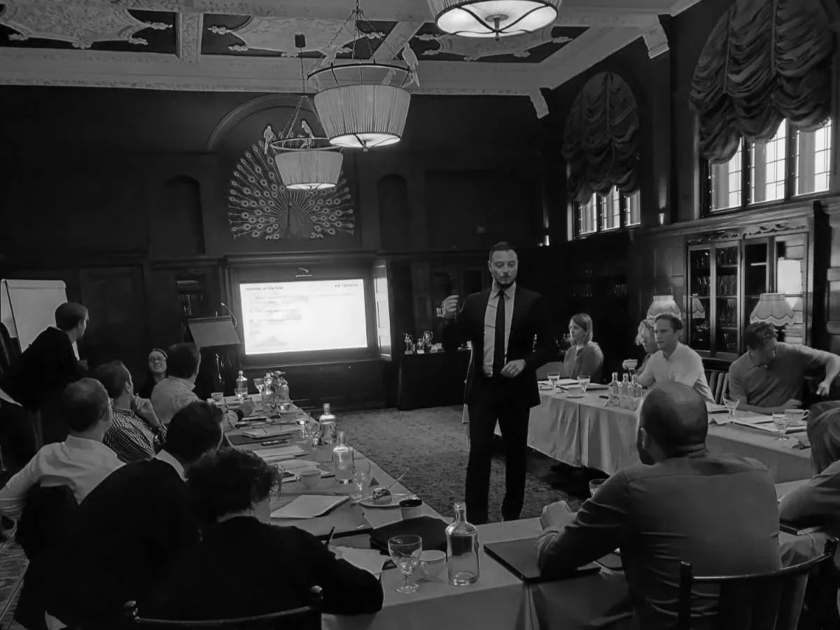 A man in a suit giving a presentation to a group of people in a formal conference room with wood-paneled walls and large windows.