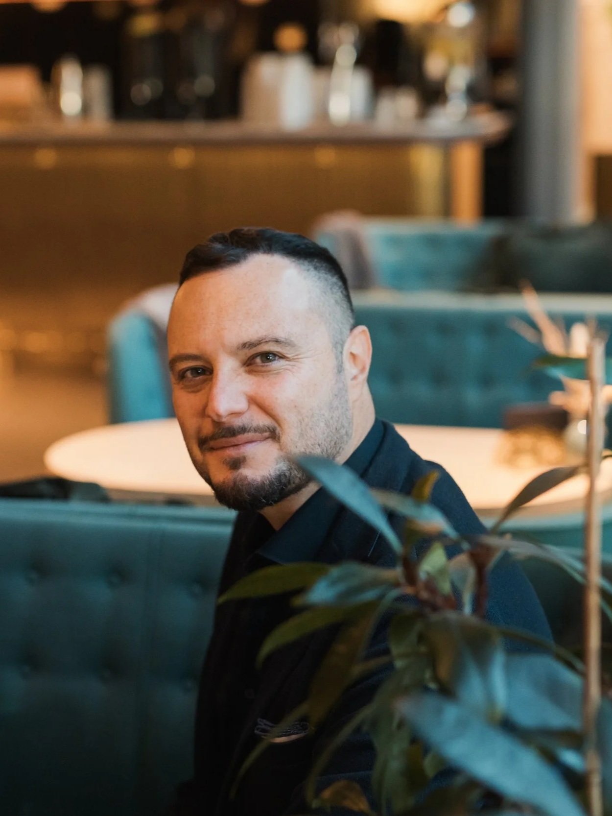 A man with short dark hair and a beard, wearing a dark shirt, sitting in a restaurant or cafe environment with blue-green seating and blurred background of tables and decor.