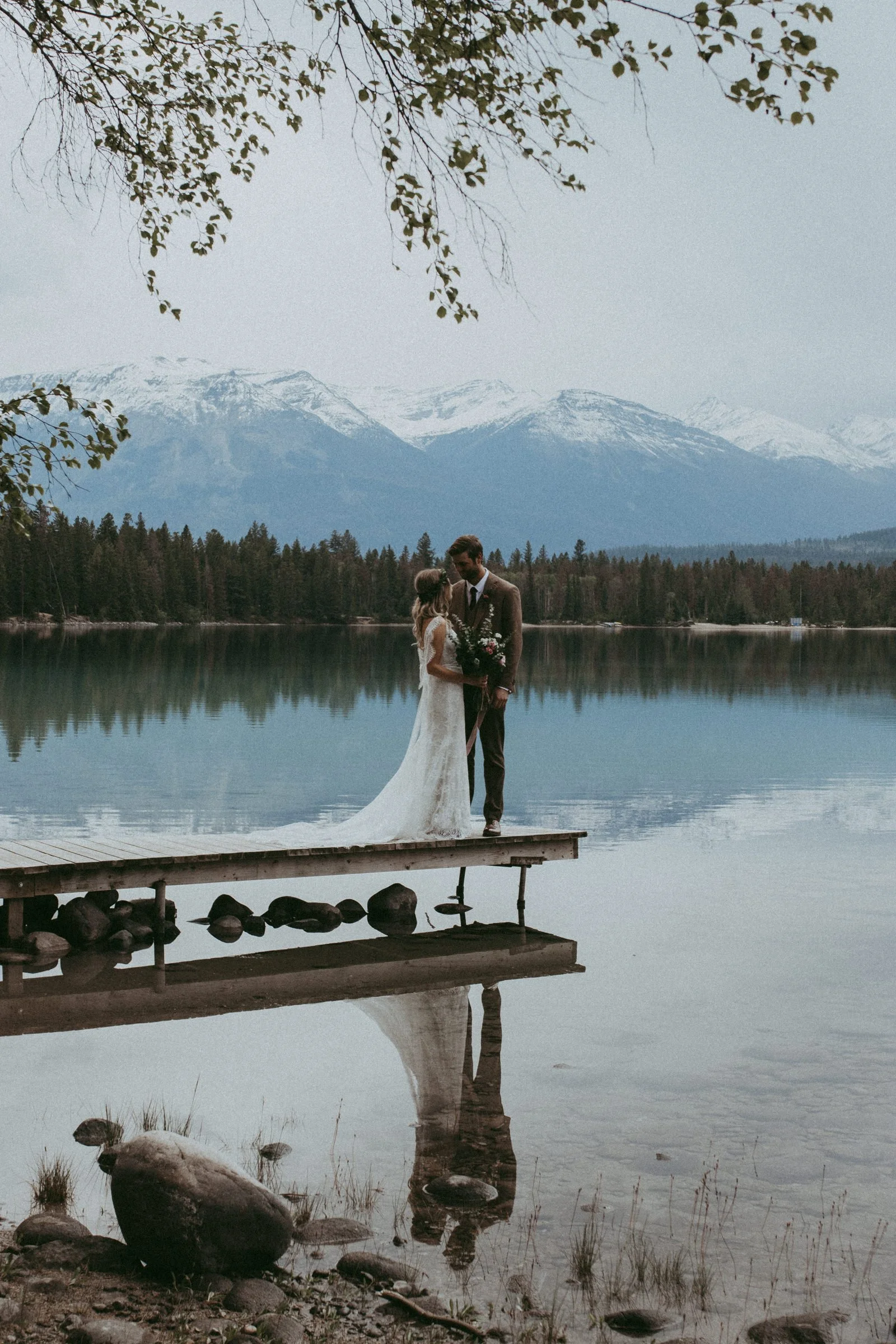 couple standing on a dock in Jasper national park during their small wedding day with an overcast sky.