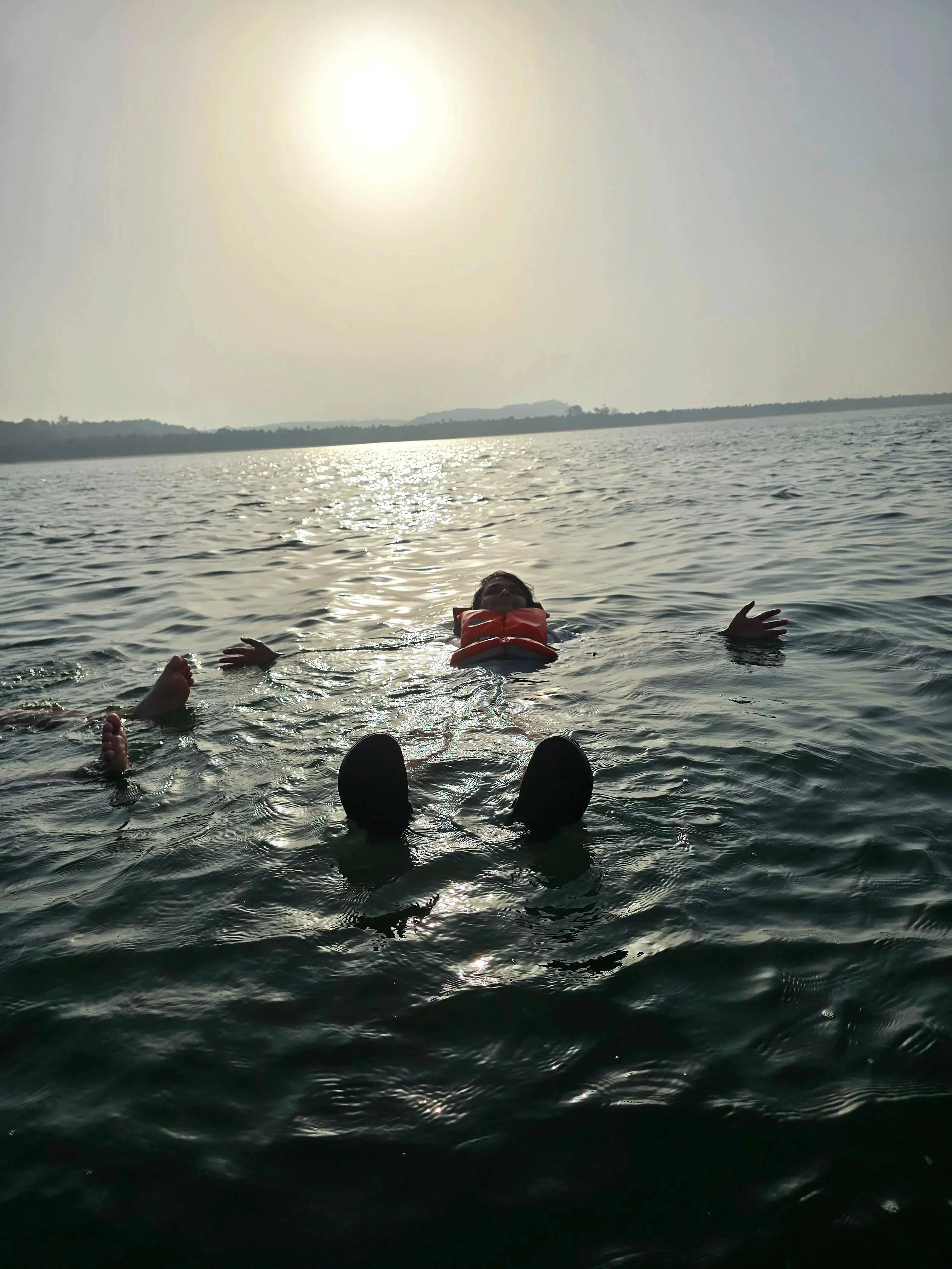 Person floating in a body of water with a life jacket in the evening, with the sun low in the sky and distant land on the horizon.