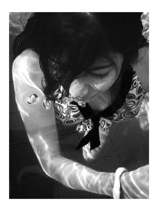 A black and white photo of a woman with dark hair, wearing a patterned top, looking down. She appears to be stretching or holding her arm out in front of her.