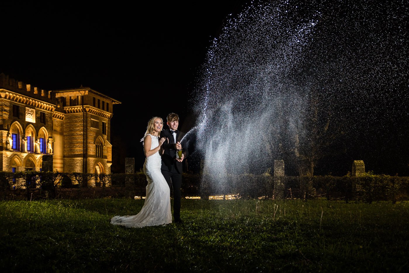 Bridal couple at night in front of wedding location with spraying champagne bottle.