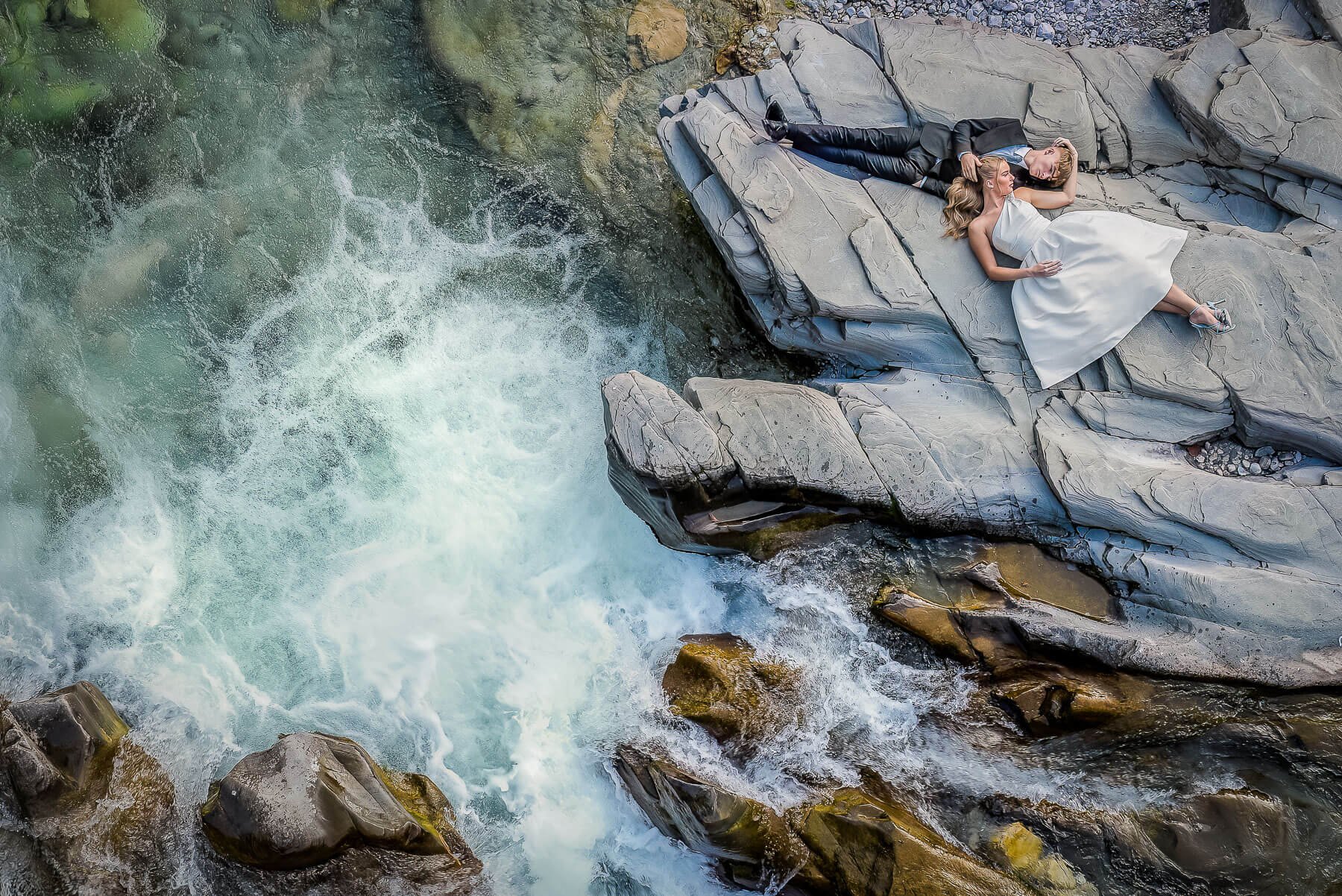 Bride and groom lying by a river on a stone ledge.