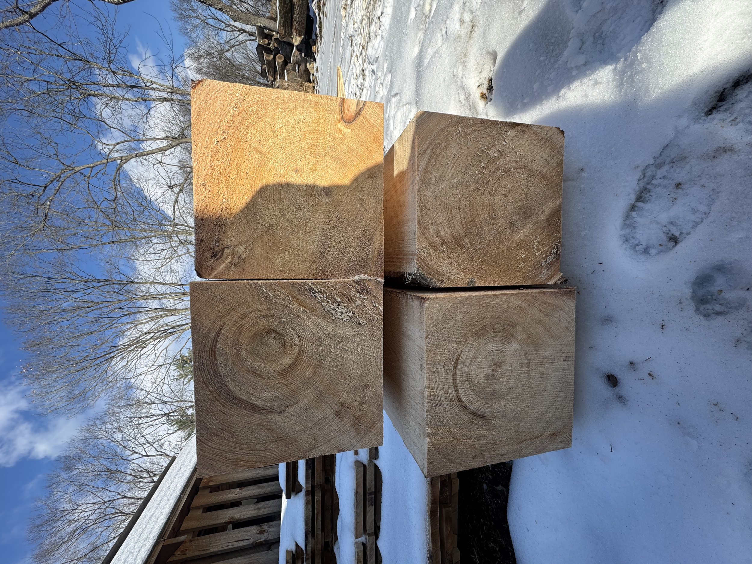 Four large wooden beams stored on snow outside, with trees, a clear sky, and a wooden fence in the background.
