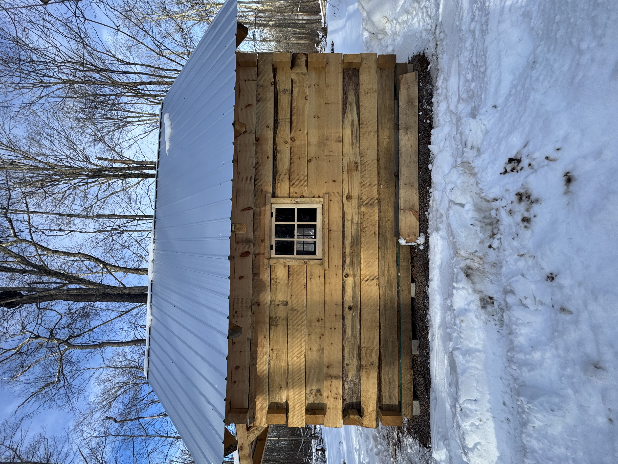 A small wooden shed with a window, built on a snowy ground outdoors, with a metal roof and leafless trees in the background.