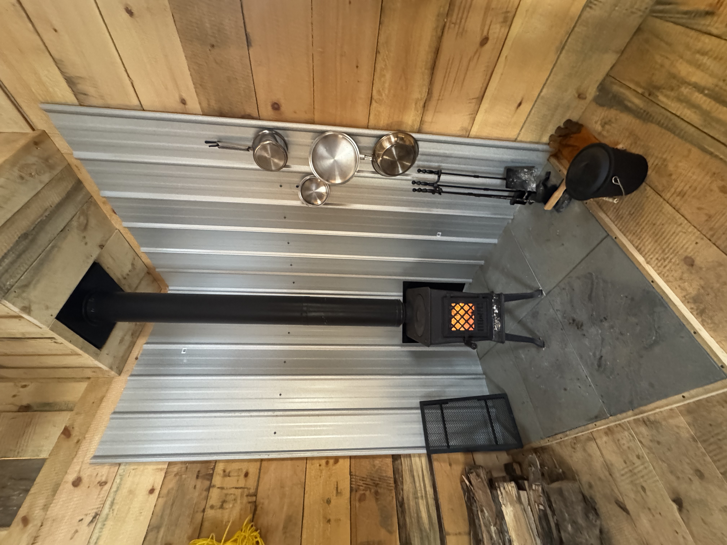 The image shows the corner of a rustic wood cabin room with a metal wall. A black stove with a pipe vent is in the corner on a concrete pad. Hanging on the wall are three metal pots and pans. Next to the stove are fireplace tools, a small black conta