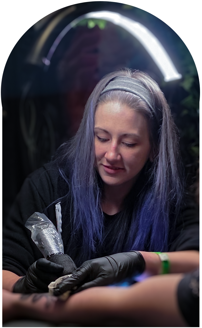 Meg Mayhem with long purple-tinted hair wearing black gloves and a headband tattooing a person's arm in a tattoo studio.