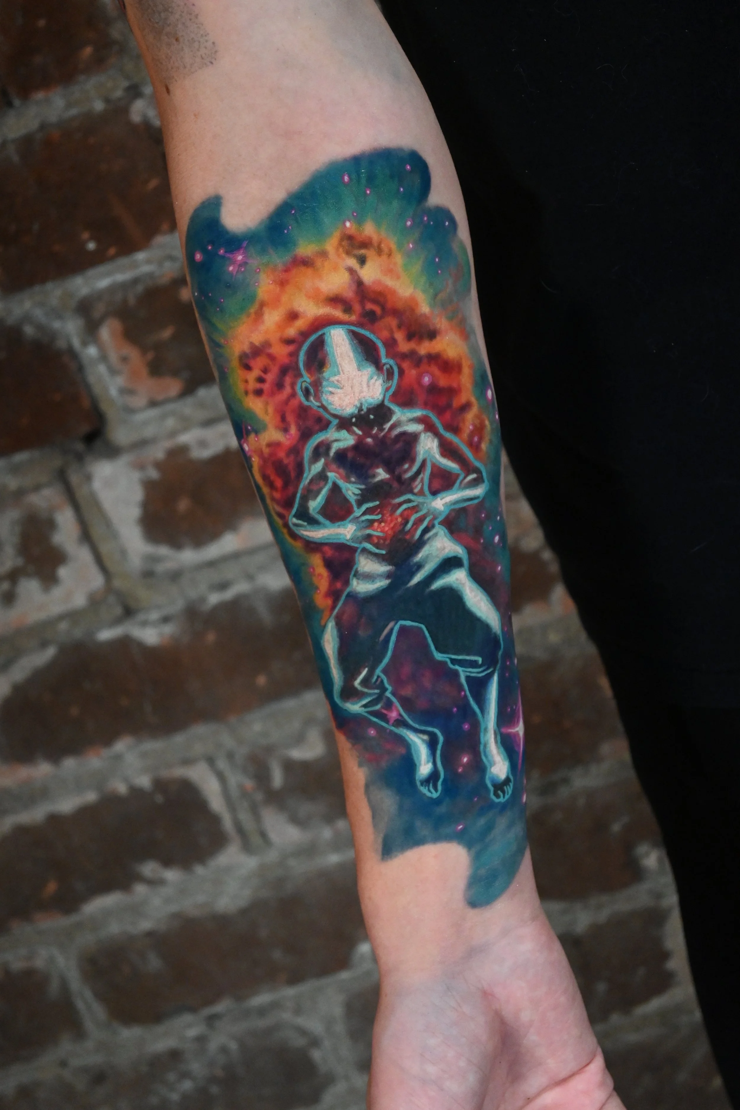 Colorful tattoo of a young boy, Aang from Avatar the Last Airbender, in a gravity-defying pose, surrounded by a cosmic, nebula background with stars and vibrant colors.