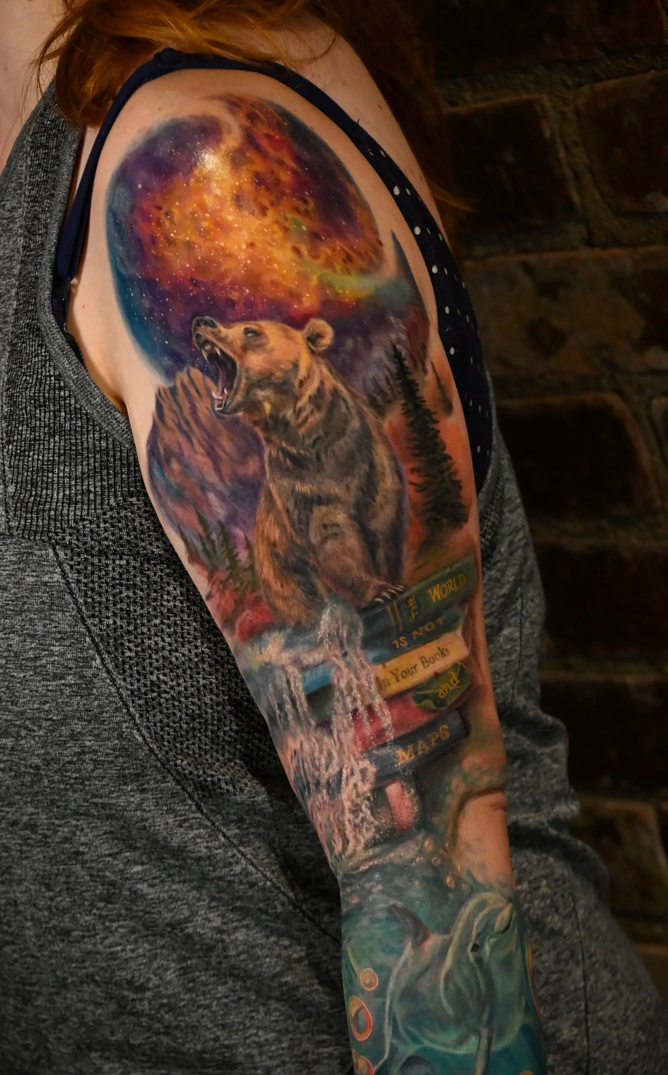 Colorful tattoo featuring a howling bear at night with a cosmic sky, mountains, trees, books, a waterfall, and dolphins.