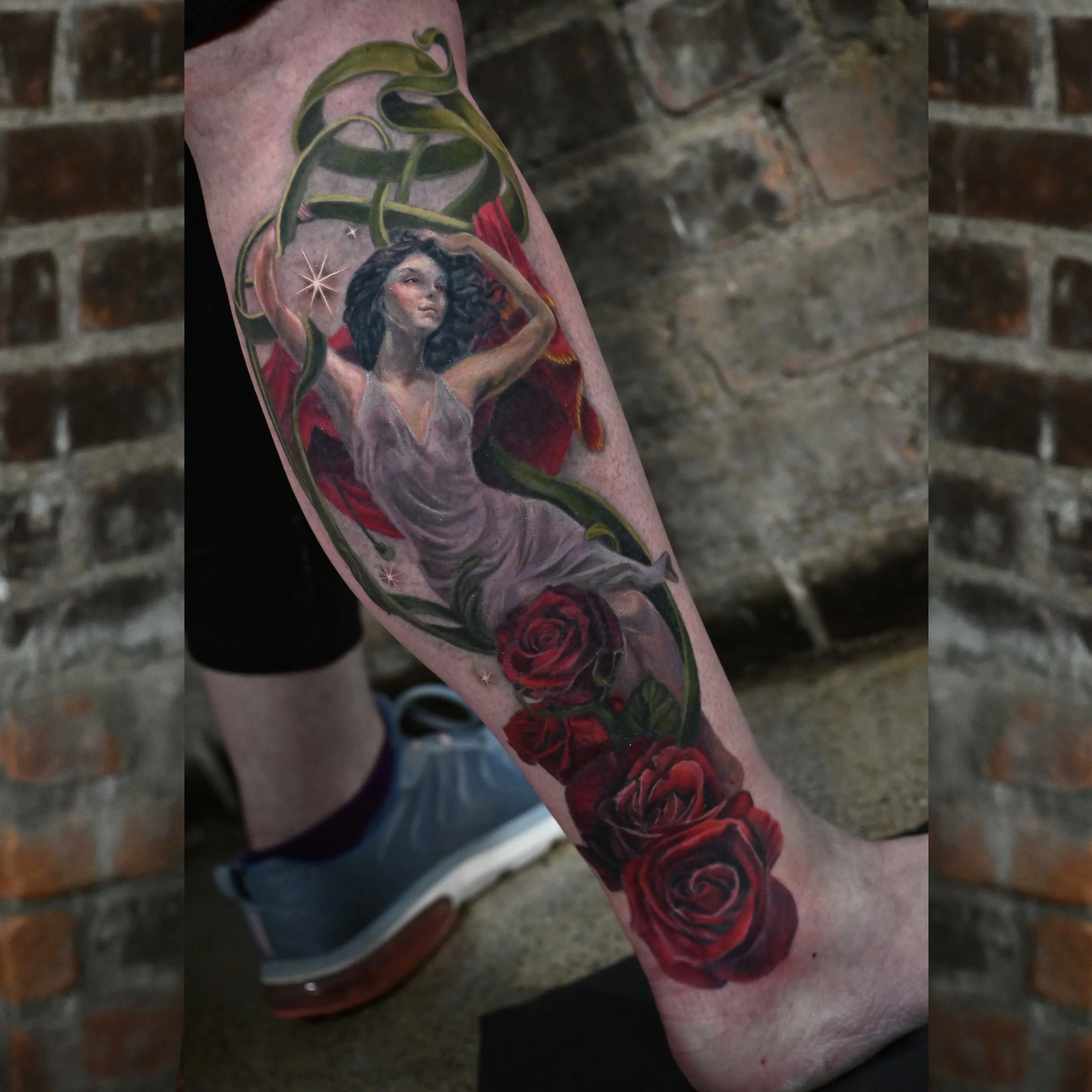 A colorful tattoo on a person's forearm featuring a woman with black curly hair, wearing a gray dress, surrounded by red roses and green vines with a brick wall in the background.