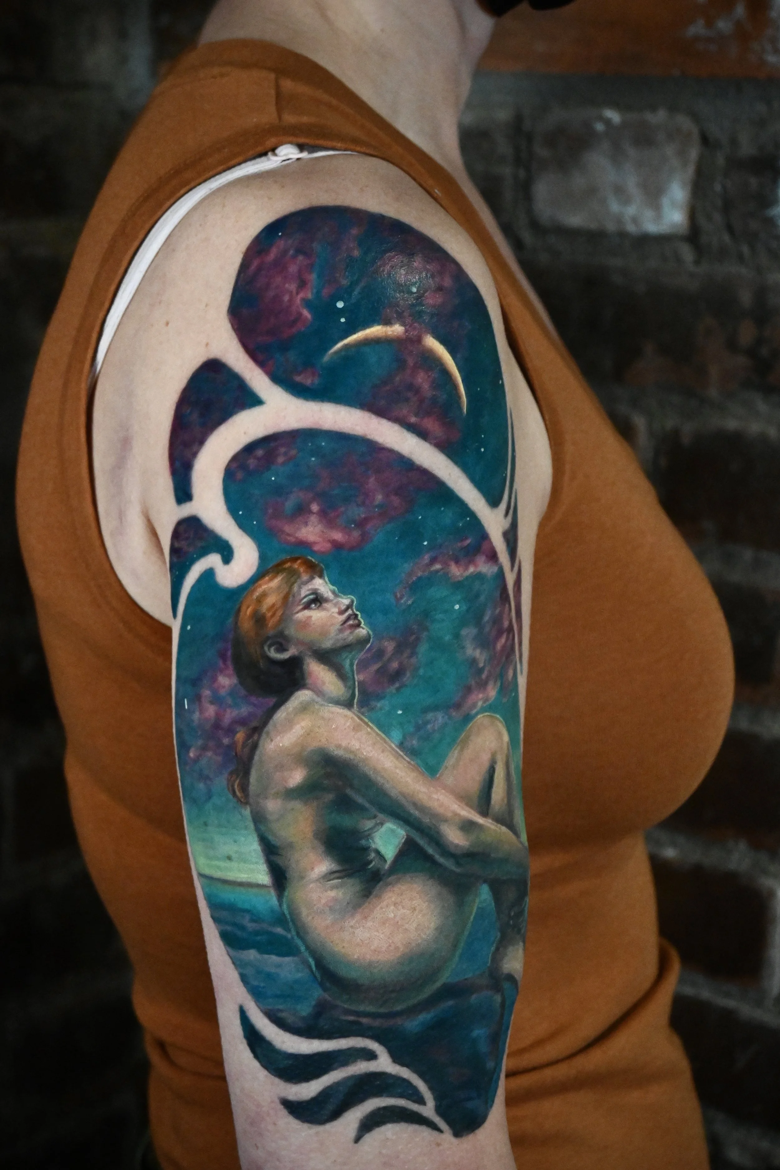 Colorful tattoo of a woman sitting by the water under a starry sky with a moon and swirling clouds on an arm.