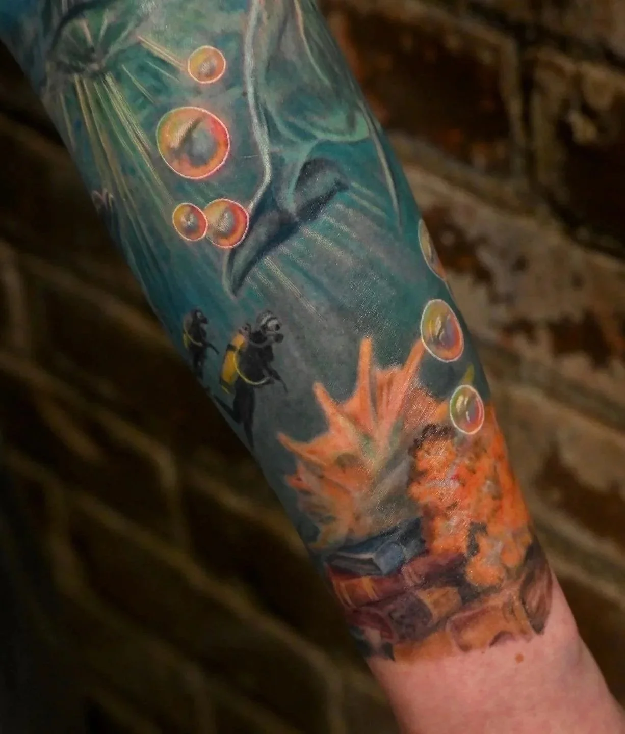 A close-up of a colorful tattoo on a person's arm, featuring several scuba divers swimming near coral and bubbles.