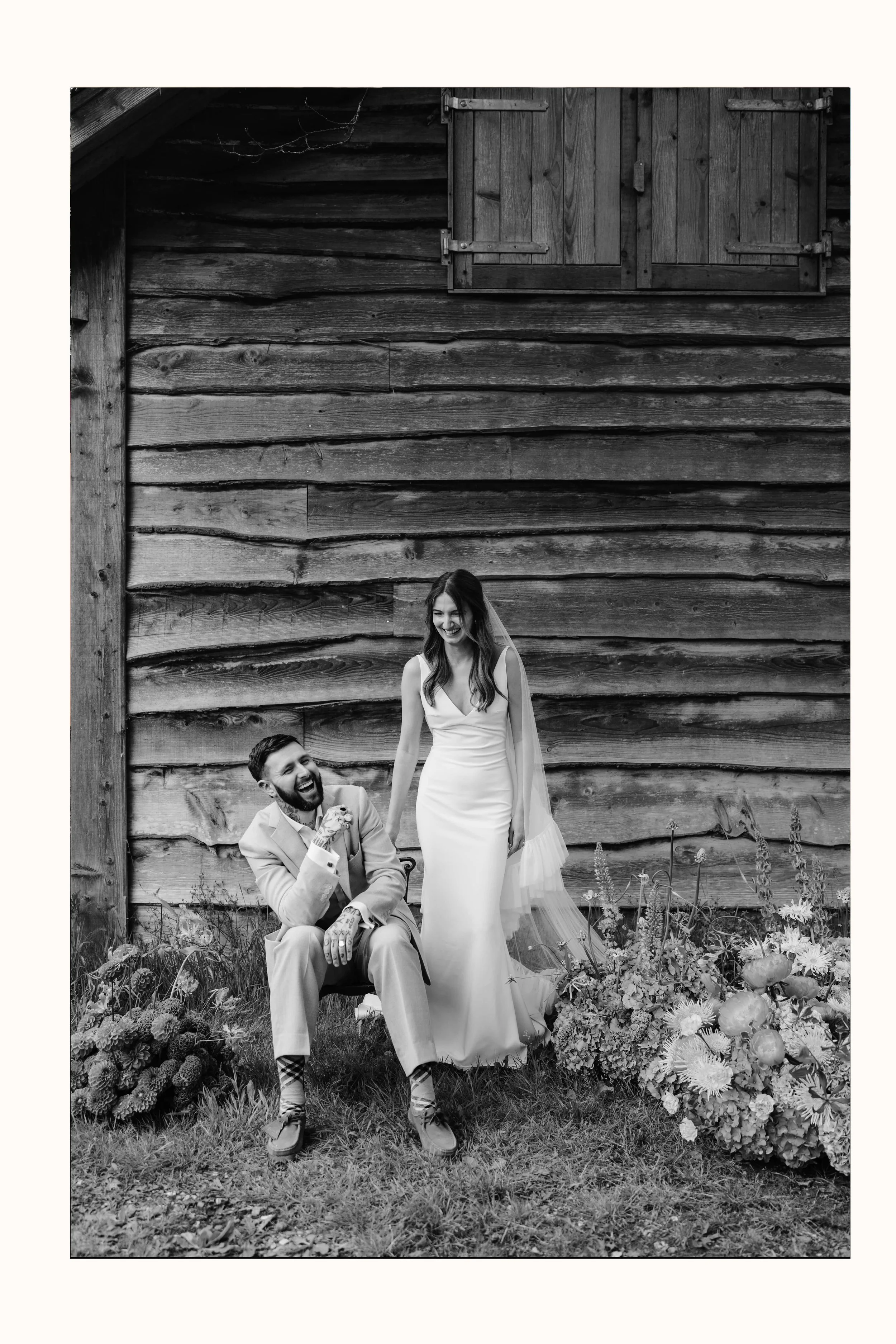 A black-and-white photo of a bride and groom laughing surrounded by flowers stood in front of building.