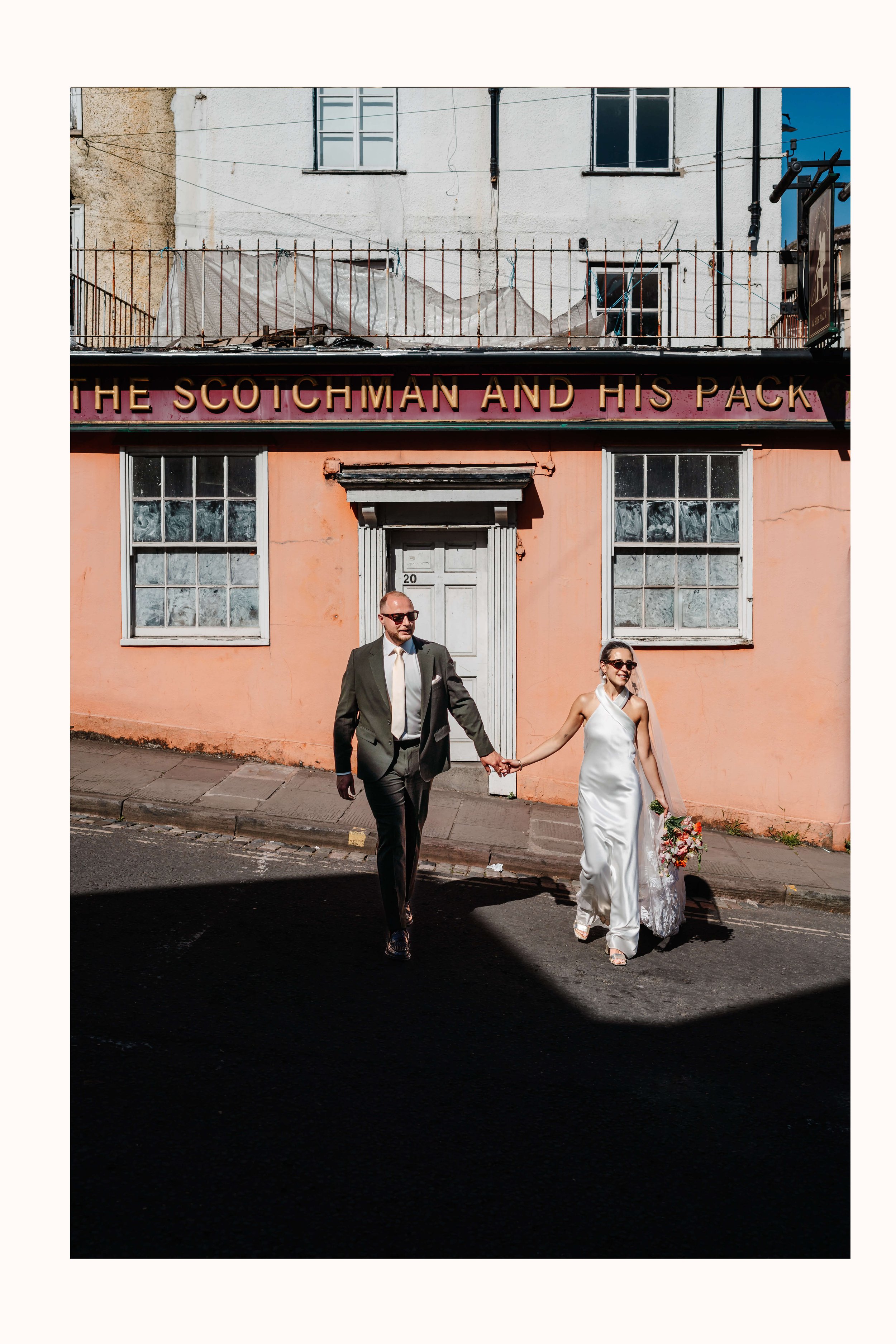 A bride and groom holding hands and walking across the road, heading to their wedding at the Mount Without in Bristol