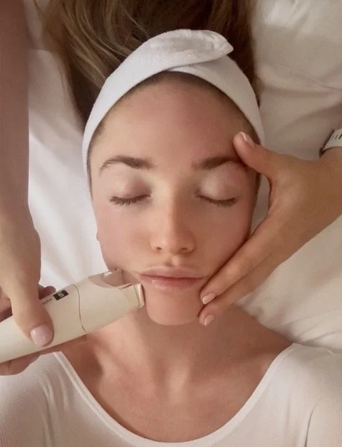 A woman with a headband is holding a young girl's face, who has her eyes closed and is receiving a facial treatment with a small device.