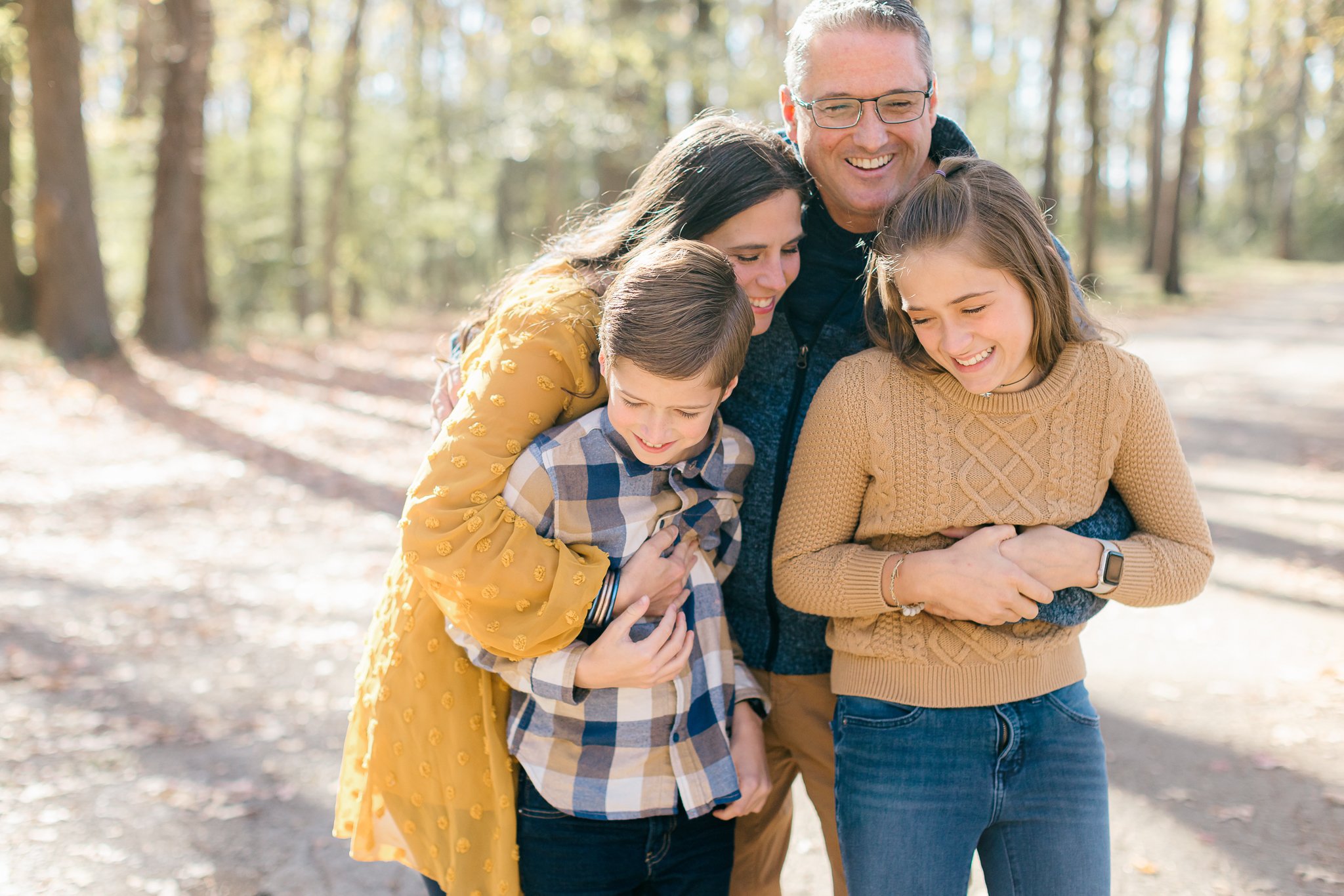 A family during a family photo session, including two children and three adults, gathering and laughing outdoors in a forested area during daytime.