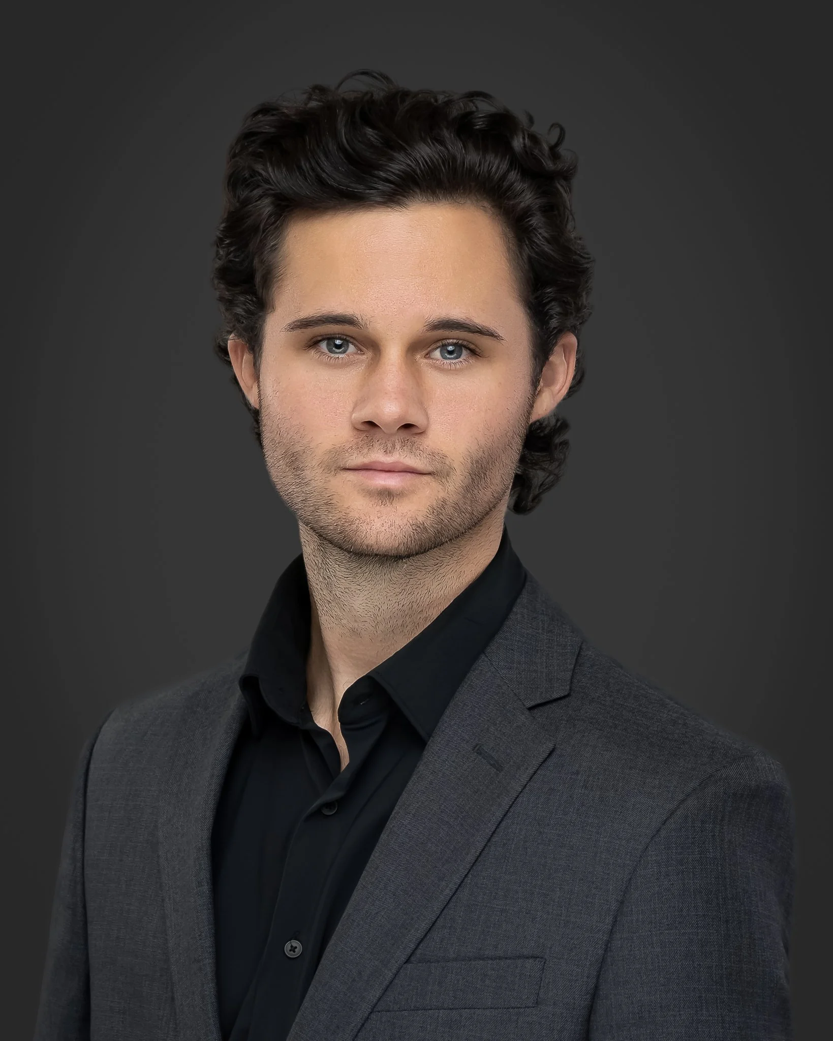 Close-up of a young man with wavy dark hair, blue eyes, and light skin, wearing a dark suit and black shirt against a dark background.