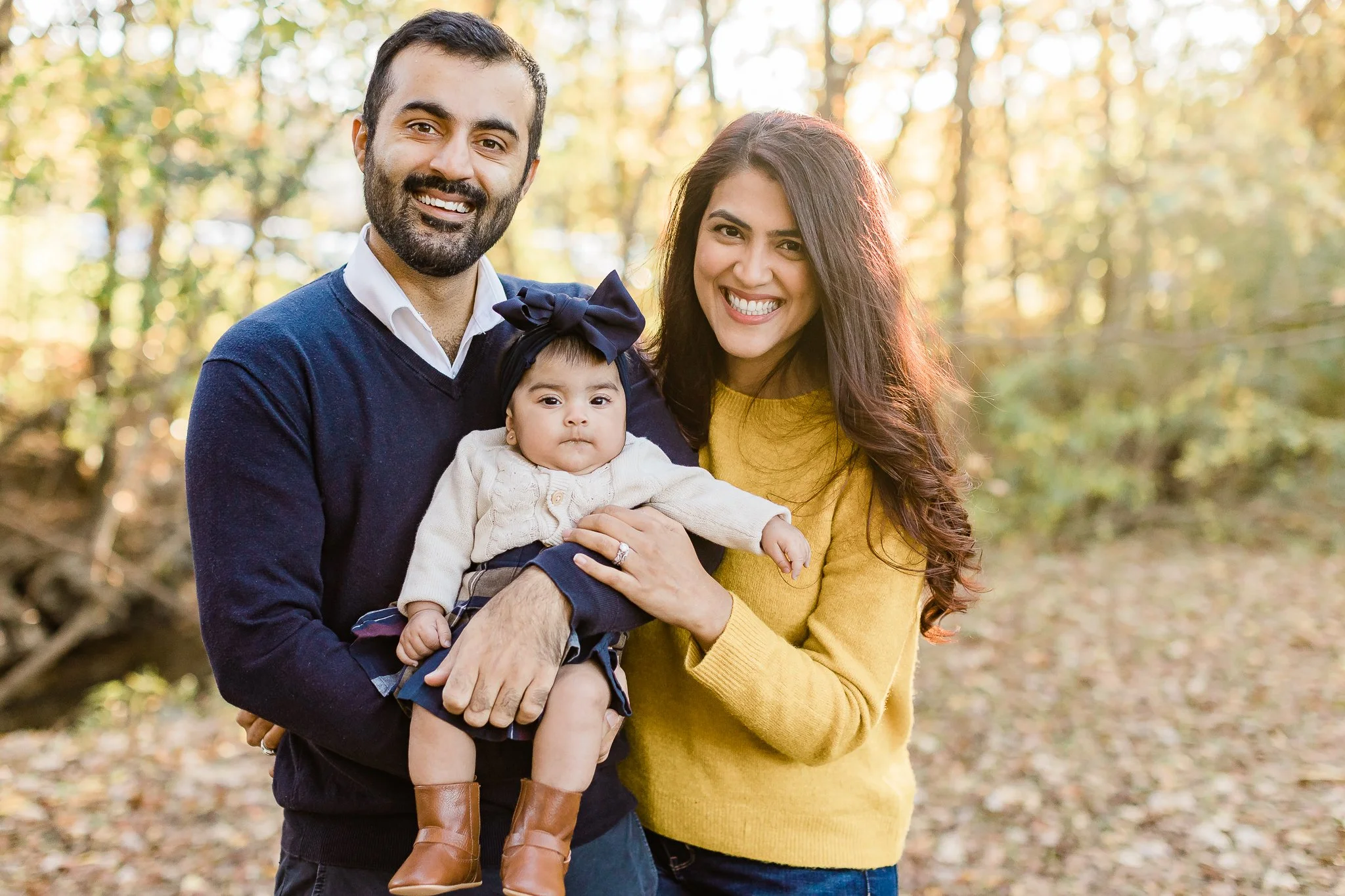 A smiling couple holding a baby in a park during fall, with autumn leaves on the ground.