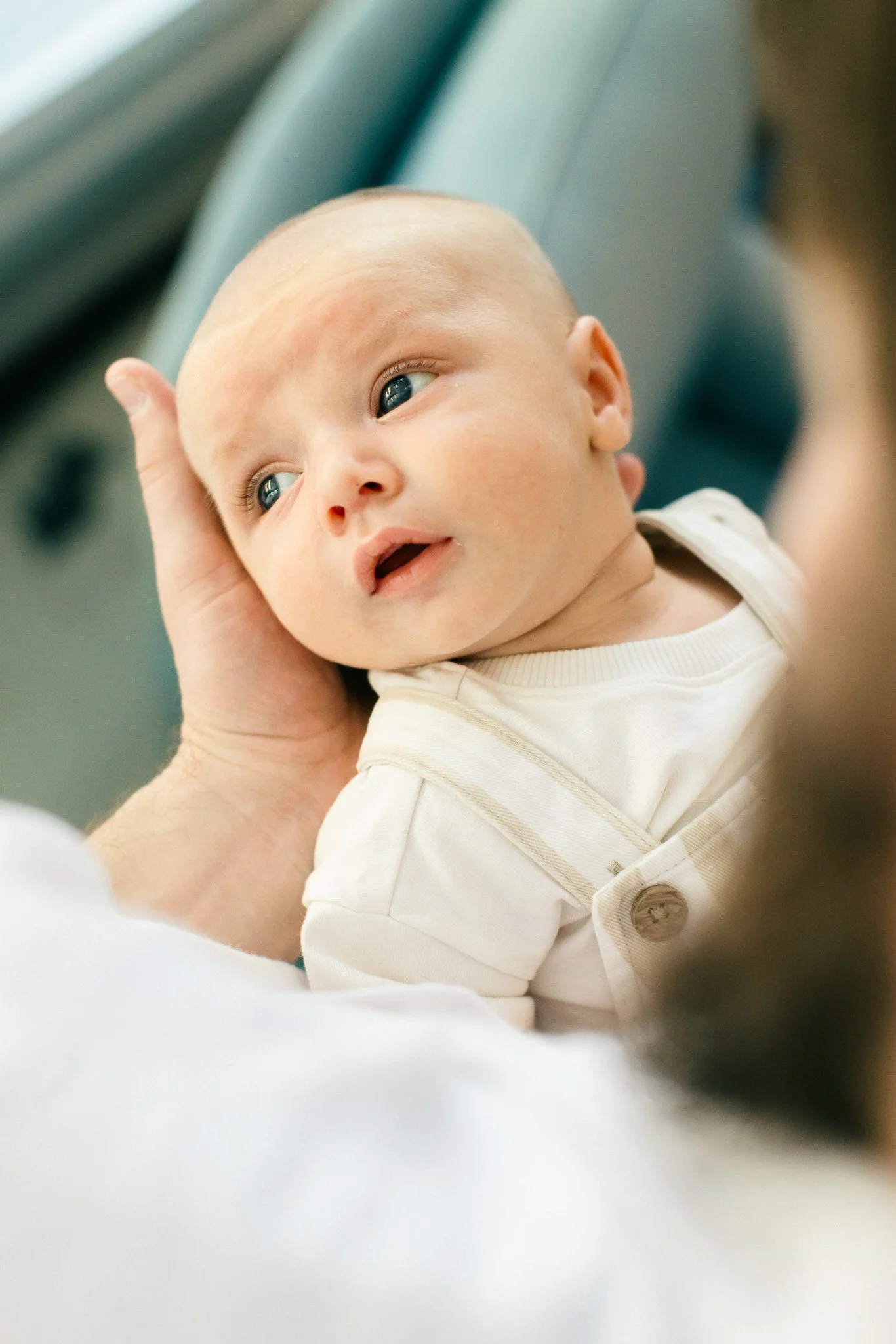 Close-up of a baby with blue eyes and a light complexion, lying on a soft surface, being gently supported by an adult hand, wearing a cream-colored outfit with buttons.