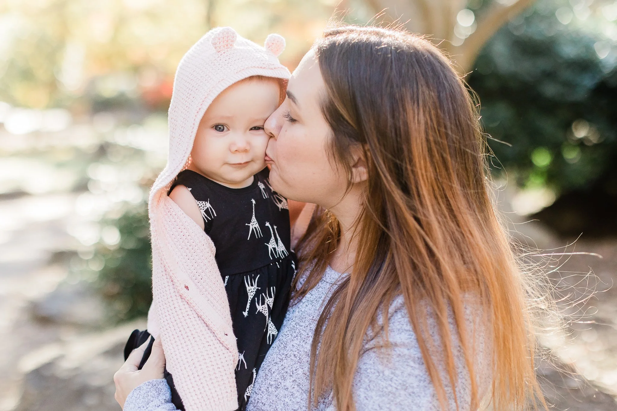 Woman kissing a baby in a pink hooded sweater outdoors.