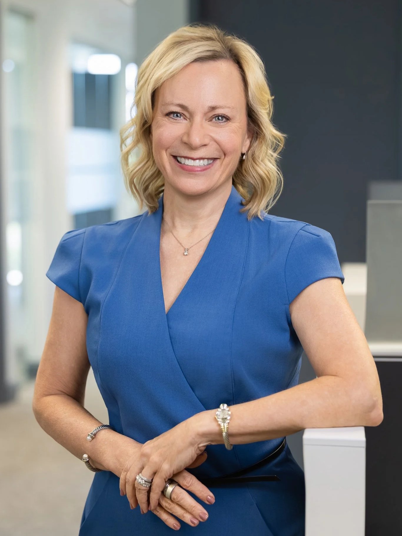 A smiling woman with wavy blonde hair wearing a blue dress with short sleeves, standing with her arms crossed in an indoor office setting.