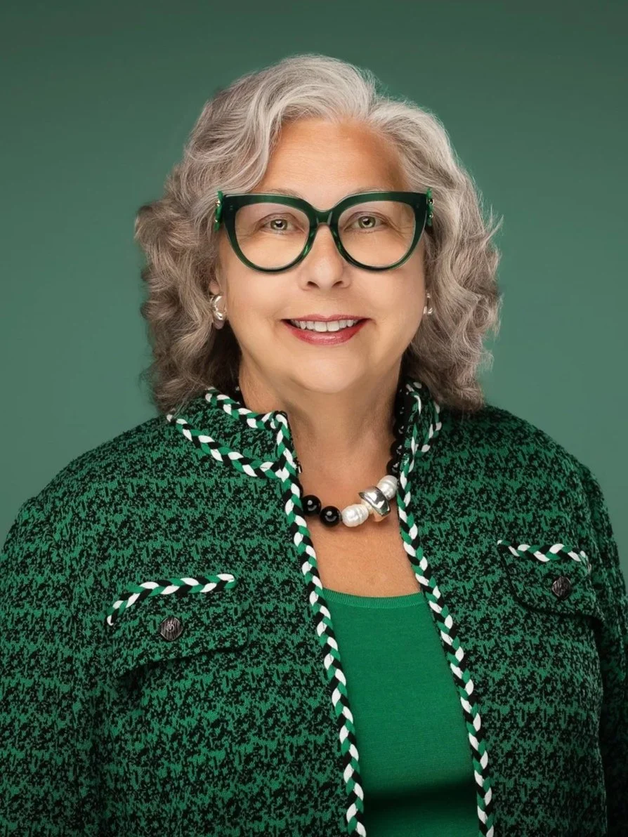 An older woman with gray curly hair, wearing glasses, a green top, and a matching green jacket with black and white details, smiling in front of a green background.