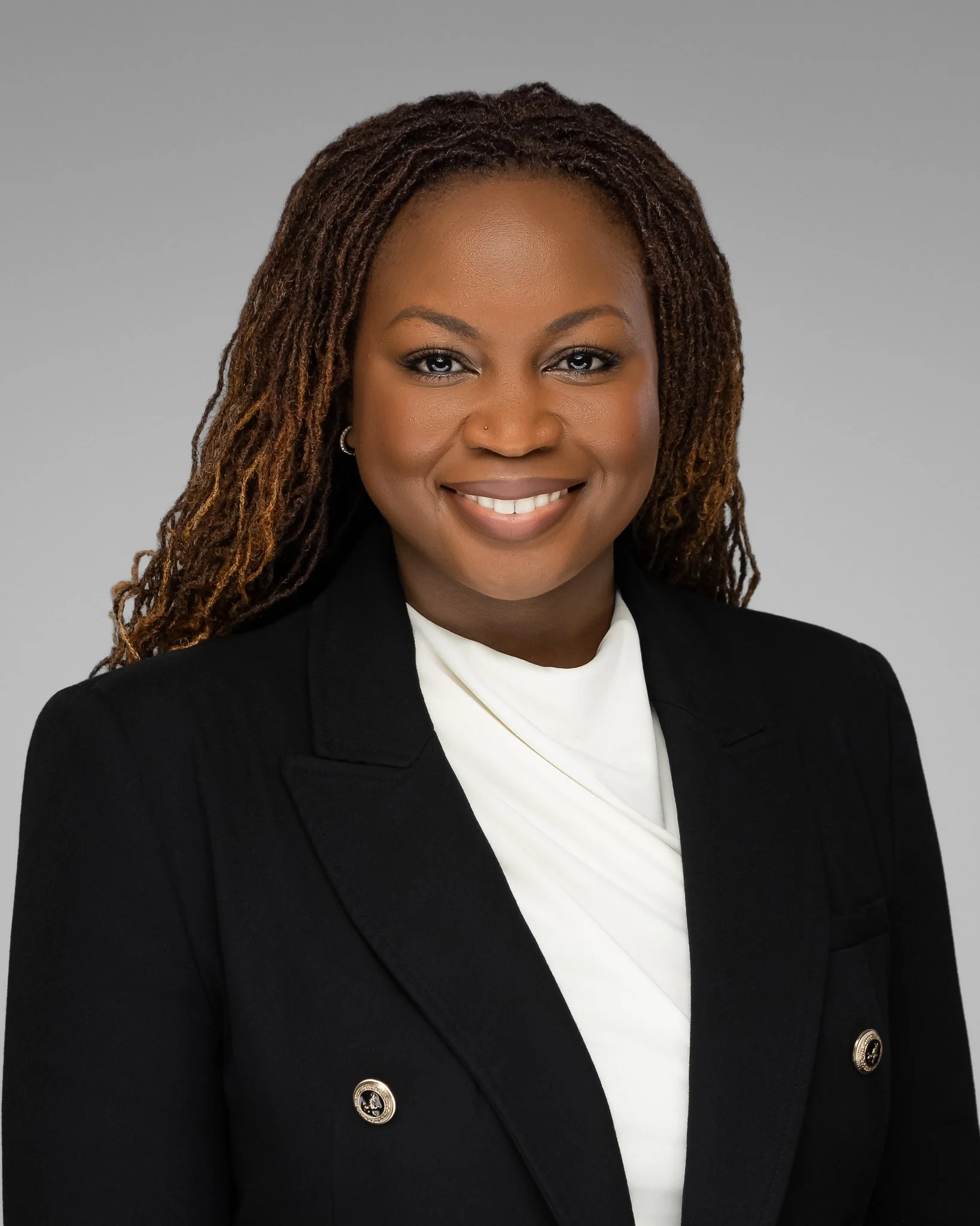 Headshot of a smiling African American woman with dreadlocks, wearing a black blazer with gold buttons and a white top, against a plain gray background.