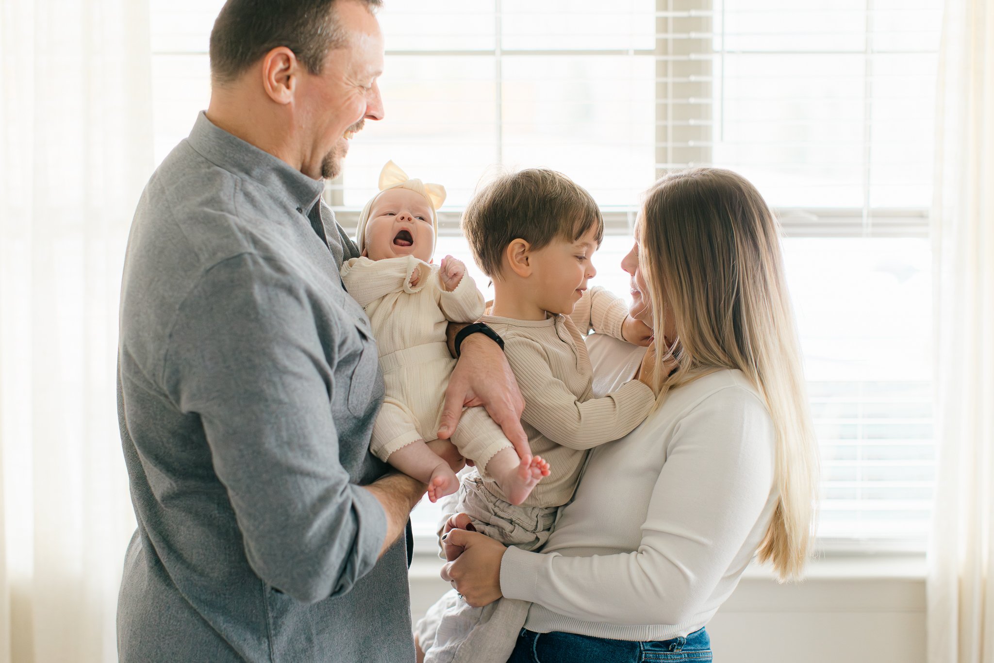 A family of four celebrating together in front of a window during a family photo session. The father holds a crying baby girl, the mother holds a smiling young boy, and they all appear happy.