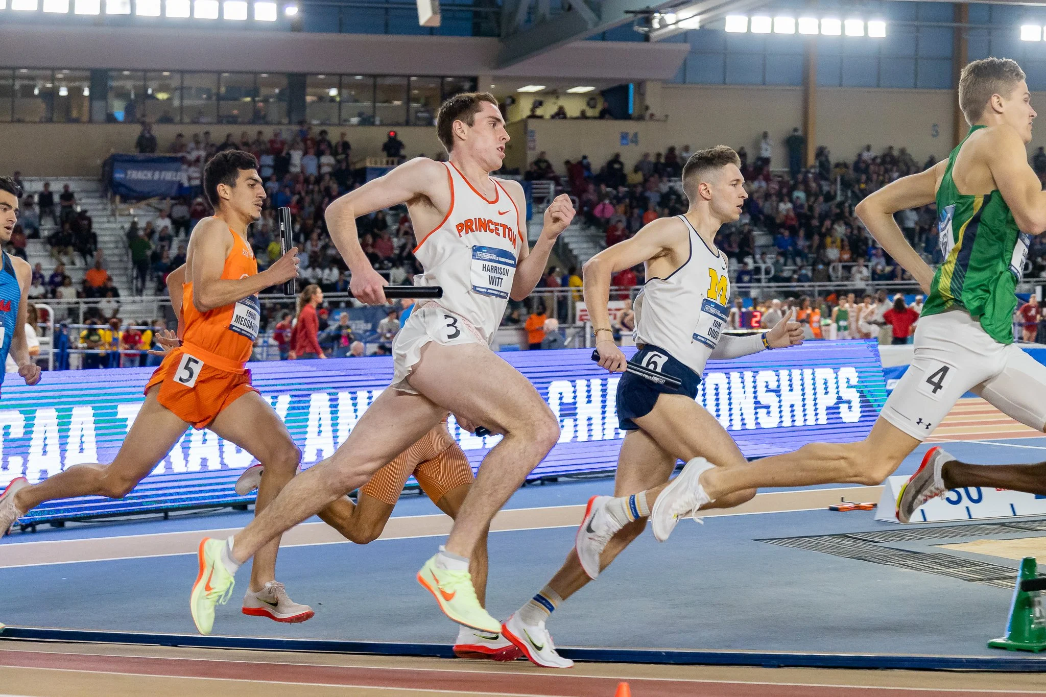 Athletes competing in an indoor track relay race, holding batons, with a crowd in attendance.