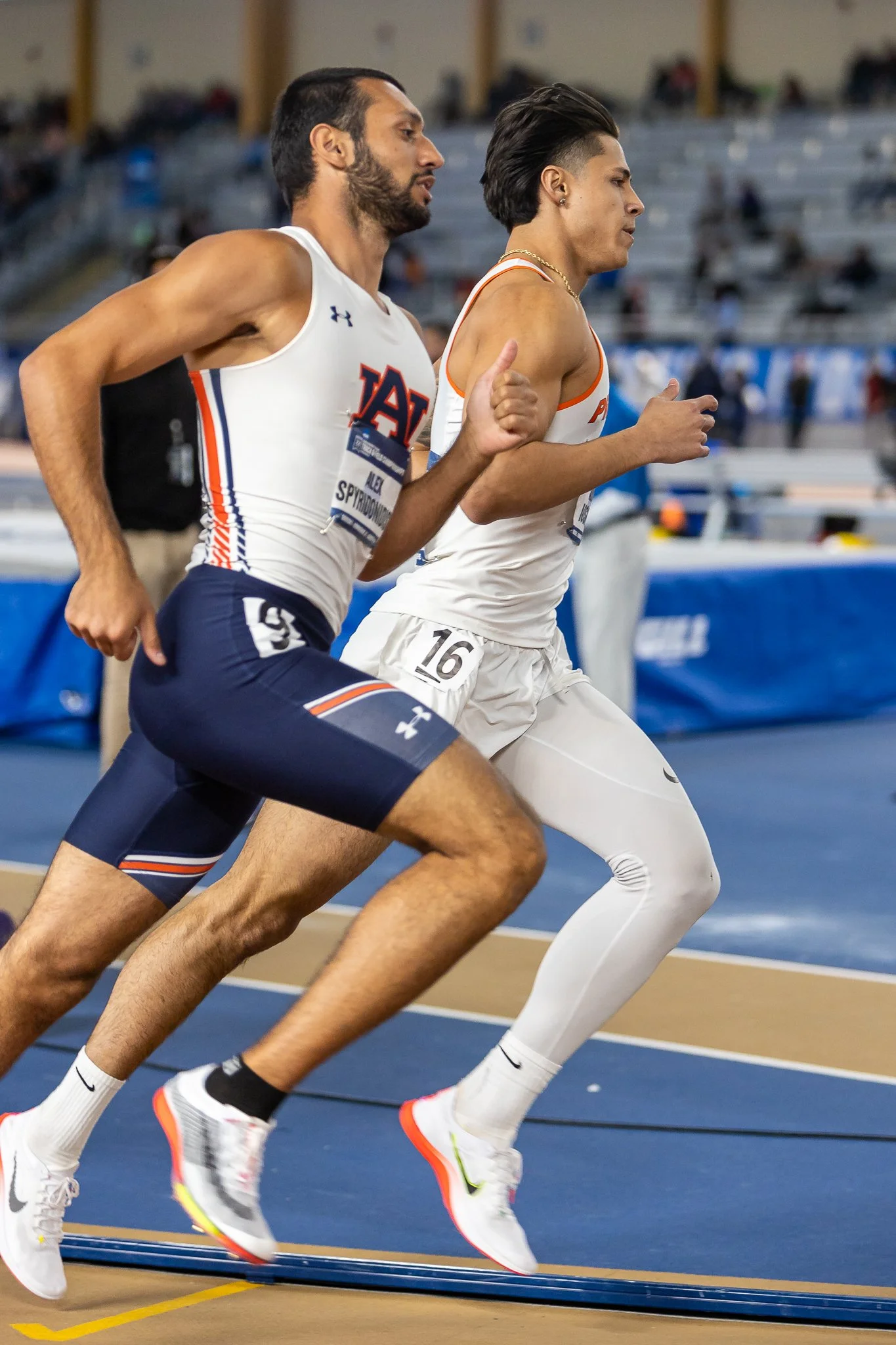 Two male athletes running on an indoor track, wearing athletic uniforms and sneakers.