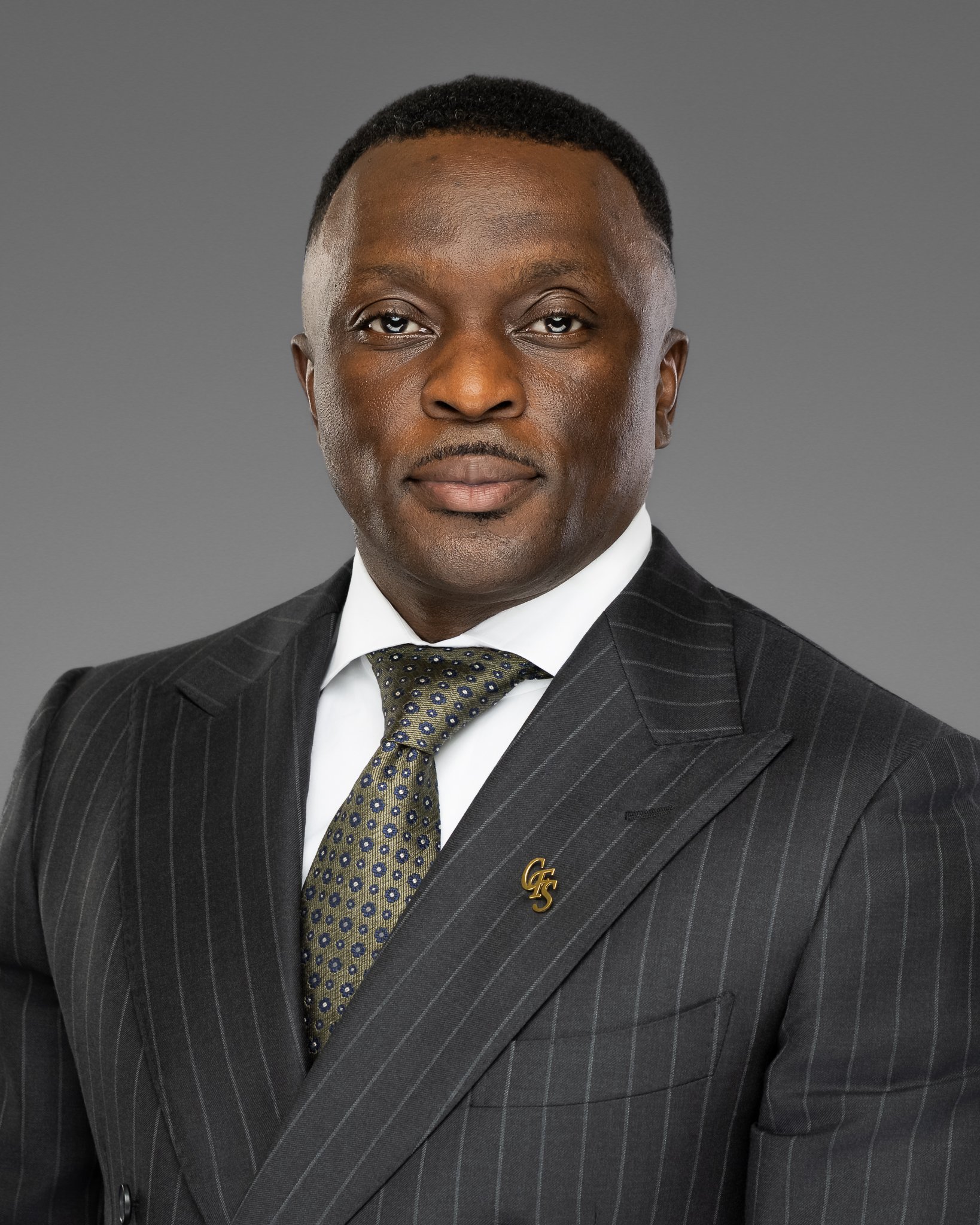 Close-up of an African American man wearing a dark pinstripe suit, white shirt, and a patterned tie, with a gold initials pin on his lapel against a gray background.