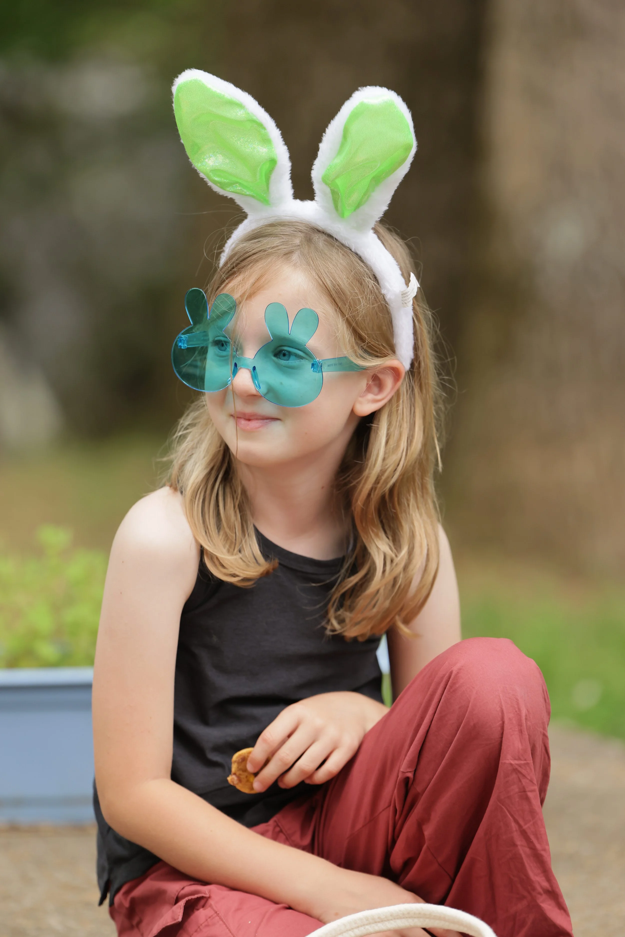 A young girl with long, blonde hair wearing bunny ears, blue rabbit-shaped sunglasses, a sleeveless black top, and red pants, sitting outdoors in a park.