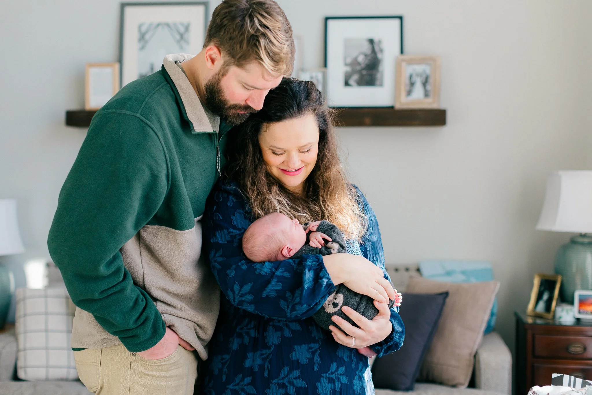A couple standing closely, with the woman holding a newborn baby. The man is wearing a green and beige fleece, and the woman is in a navy blue dress. They are in a cozy living room with picture frames on the wall.