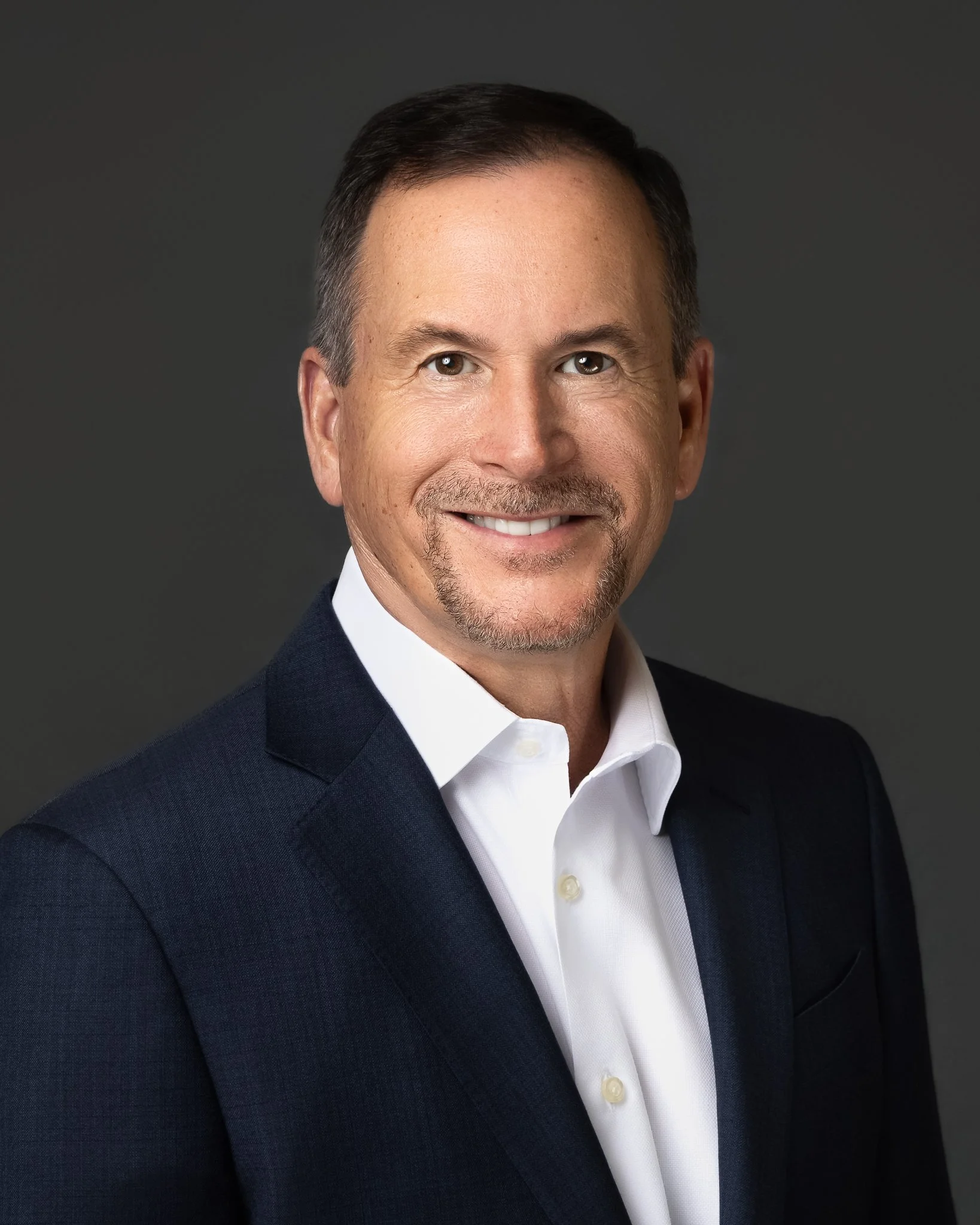 Close-up portrait of a smiling middle-aged man with short dark hair, wearing a dark suit jacket, white dress shirt, and no tie, against a dark gray background.