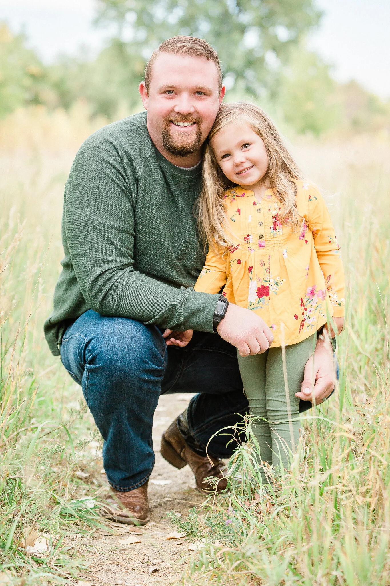 A man kneeling beside a young girl in a yellow dress, both smiling, in a grassy field.