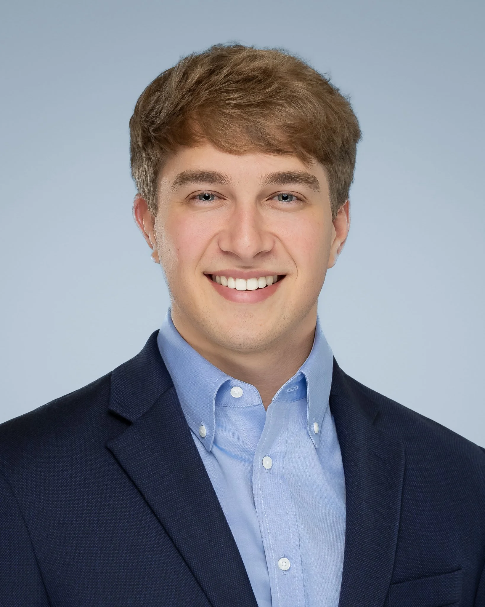 A professional headshot of a young man in a blue button-down shirt and a dark blazer, smiling against a light blue background.