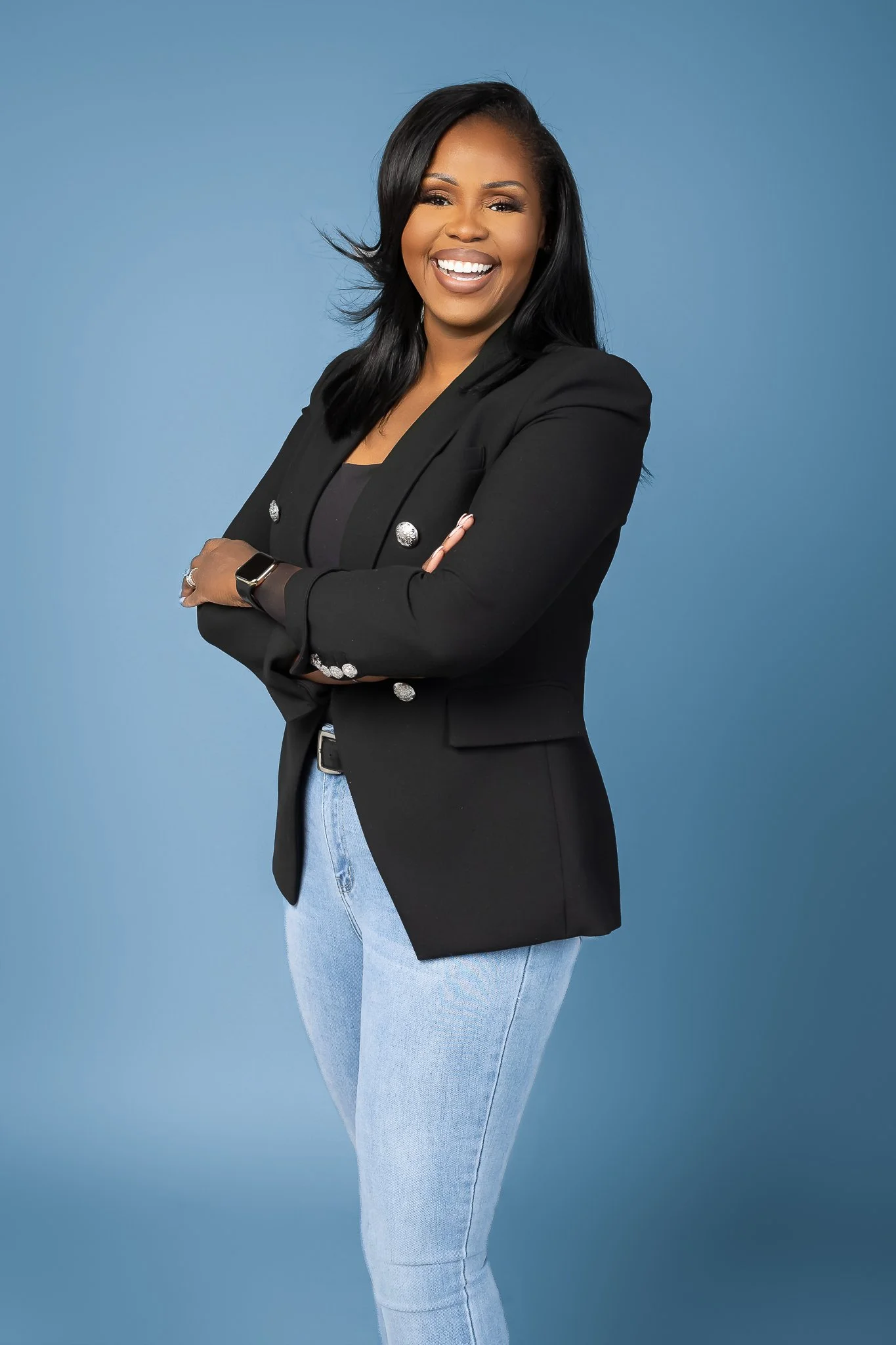 A woman with dark hair smiling, wearing a black blazer with shiny buttons and light blue jeans, standing against a blue background.