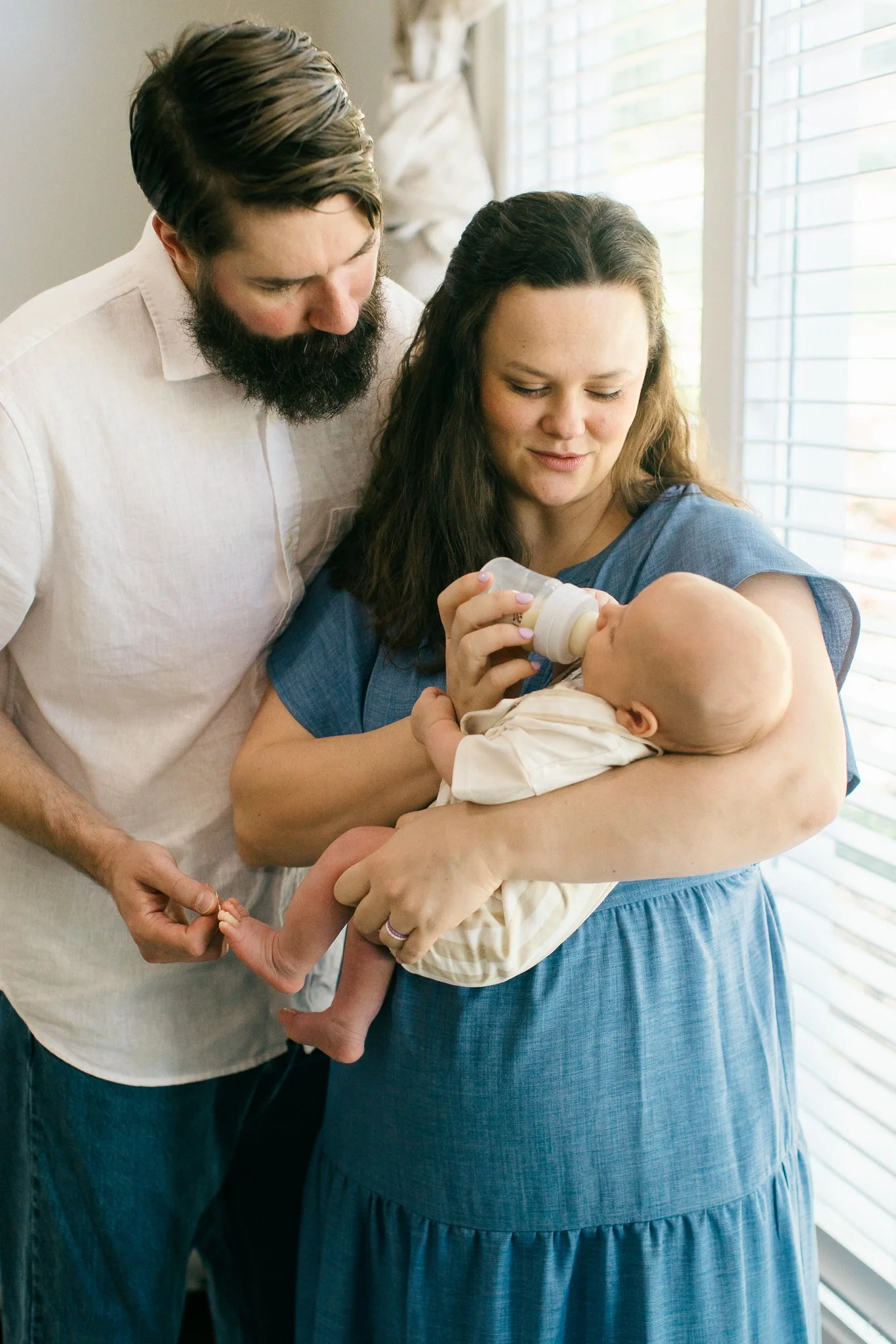 A woman holding a baby and bottle feeding it while a man looks on, inside a bright room with window blinds.