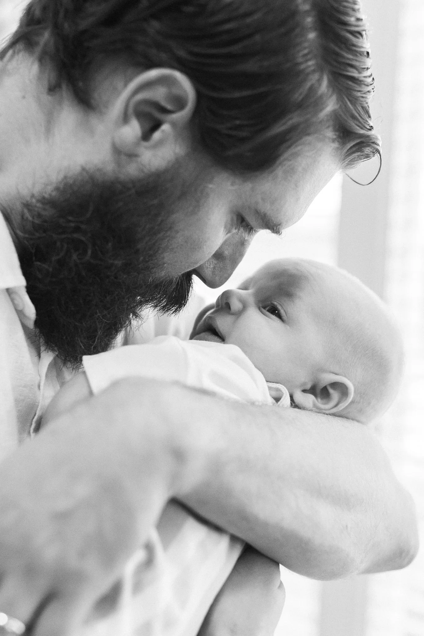 A black and white close-up photo of a man with a beard and slicked-back hair holding a baby close to his face, both looking at each other.