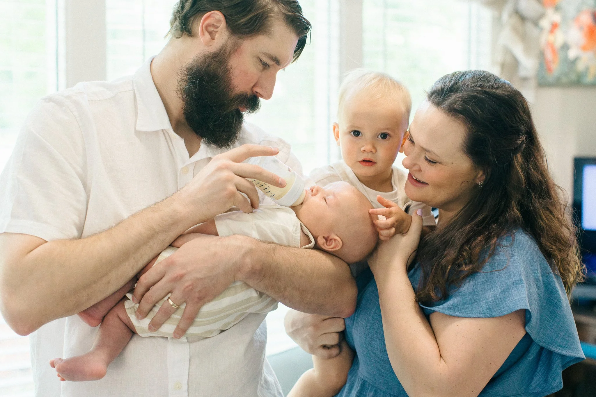 A man handing a baby bottle to a newborn baby held by a woman, with a young girl looking on, in a bright room with large windows.